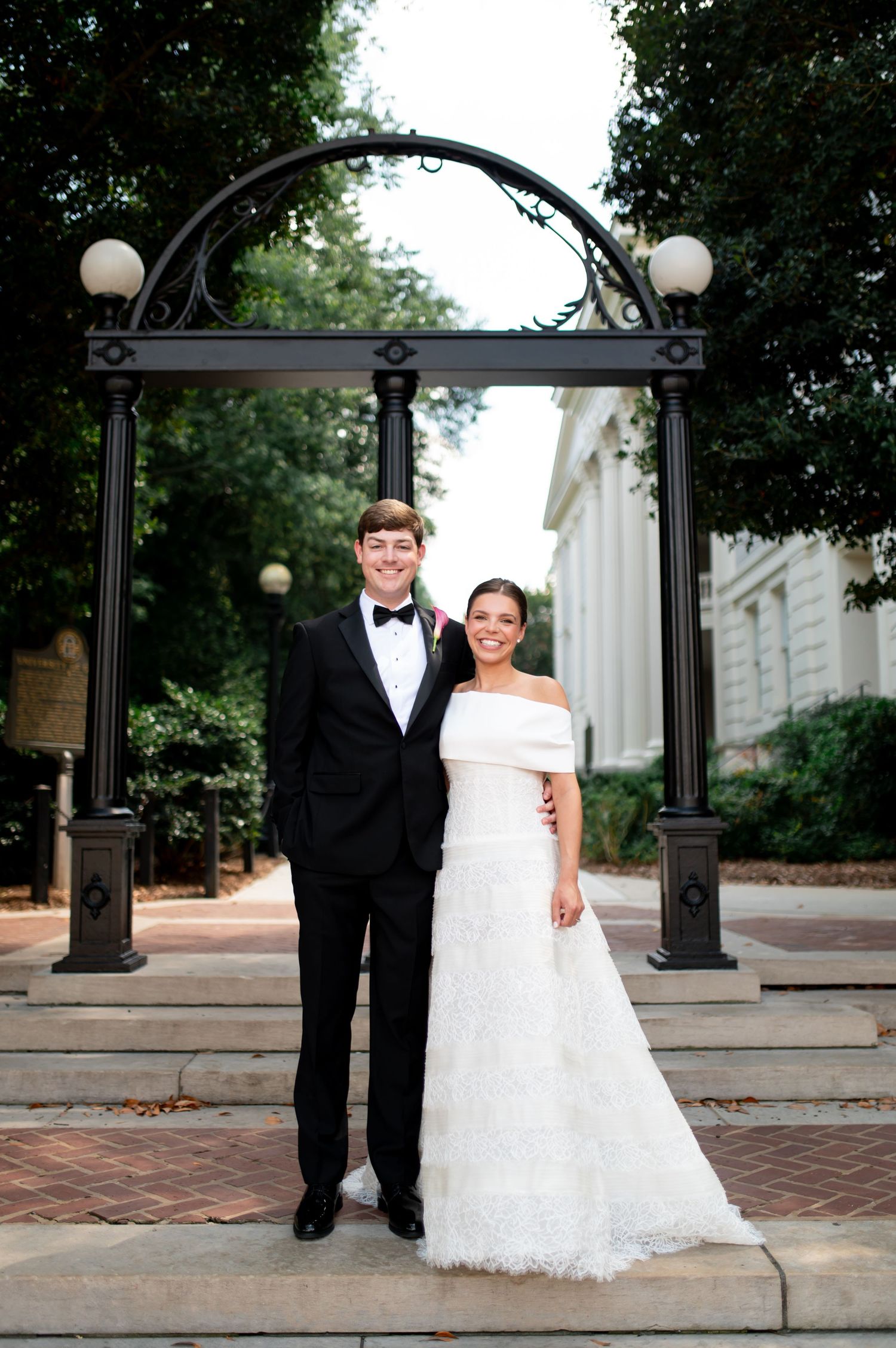 Bride and groom pose under iconic arch landmark in classic black tuxedo and off-shoulder lace wedding gown.