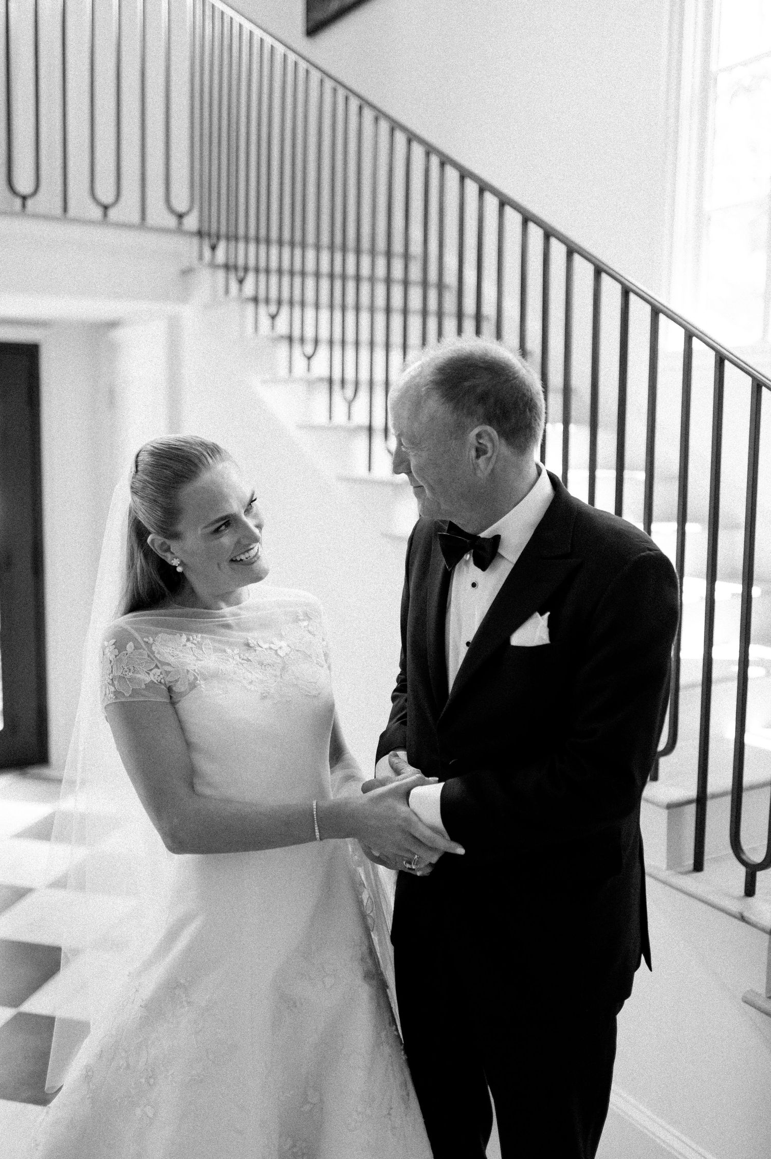 Bride and groom share emotional first look moment holding hands by staircase in elegant black and white wedding photo.