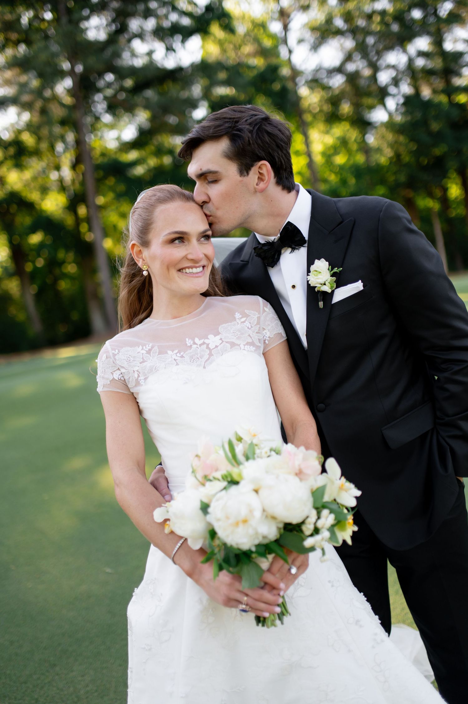 Groom in black tuxedo kisses bride wearing lace gown holding white peony bouquet.
