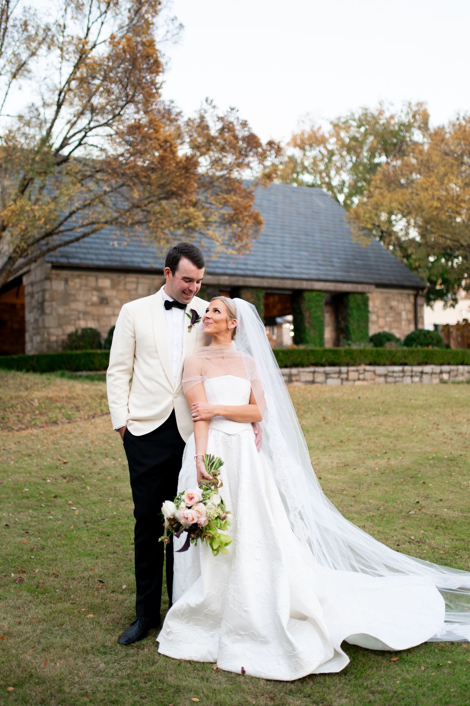 A newlywed couple takes wedding portraits on a grassy lawn with a stone building in the background.