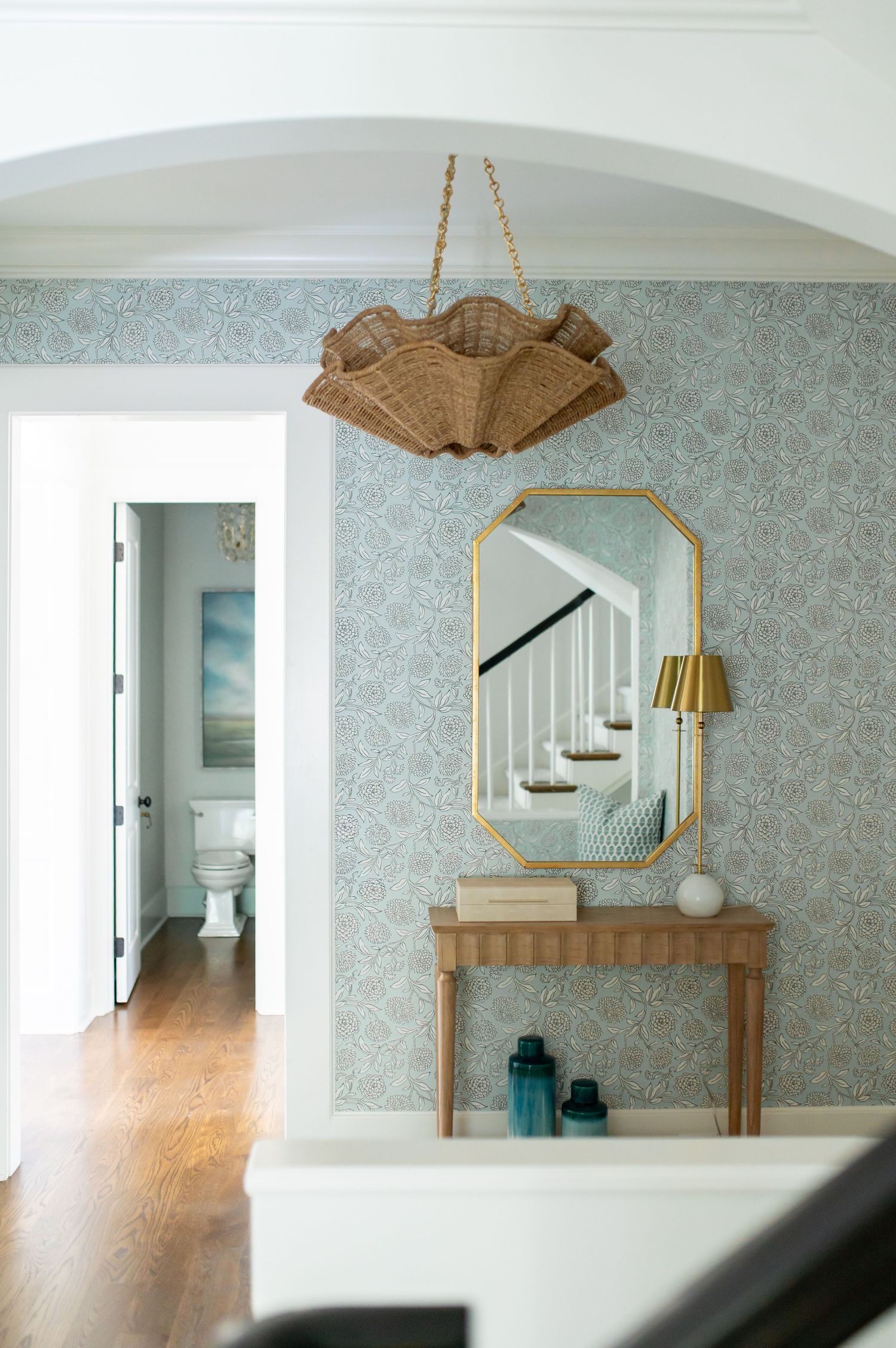 Elegant entryway with blue floral wallpaper, gold octagonal mirror, rattan chandelier, and wooden console table with teal accents.
