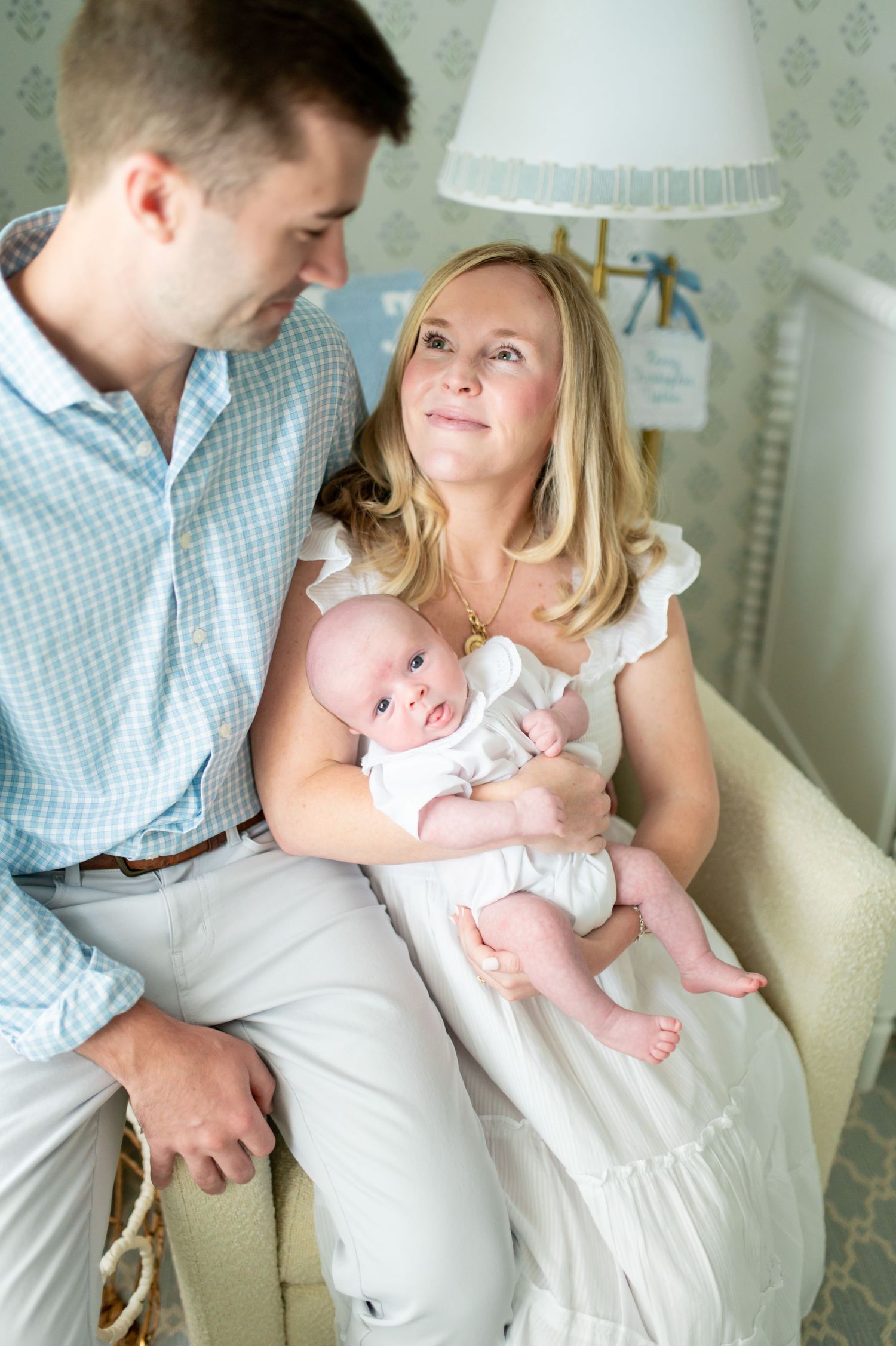 Young parents sitting together, mother holding newborn baby in white outfit, in a softly decorated nursery room.