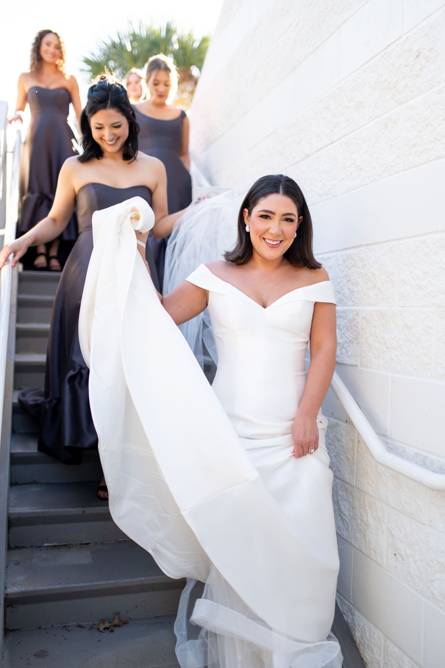 Bride in flowing white dress posing with bridesmaids against white wall.