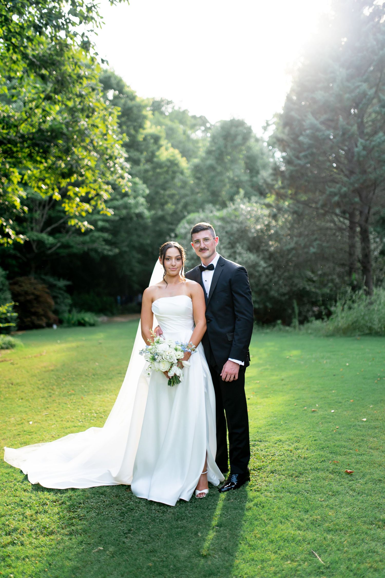Wedding couple poses together on lush green lawn during outdoor photo session.
