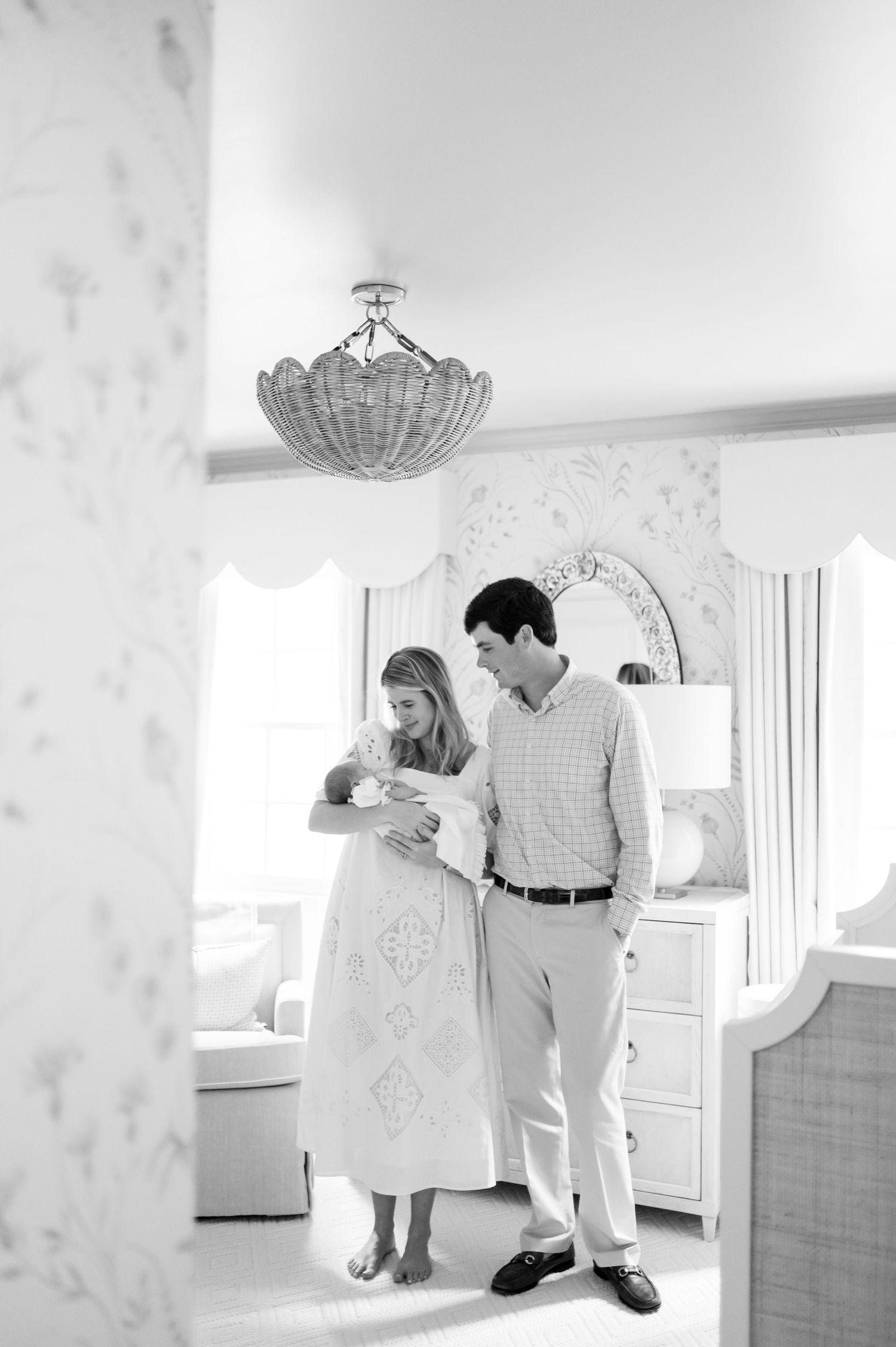 A couple stands in a bright nursery room with an elegant chandelier and floral wallpaper.