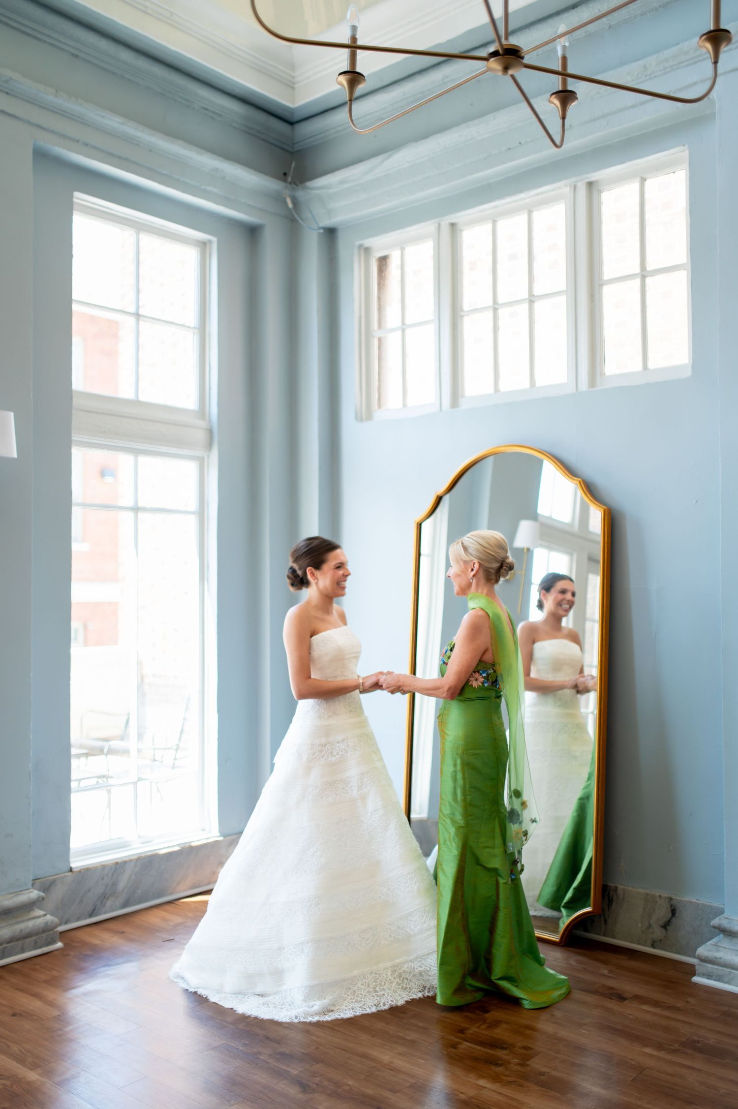 Bride in white strapless gown holds hands with woman in green dress beside gold floor mirror in blue room.