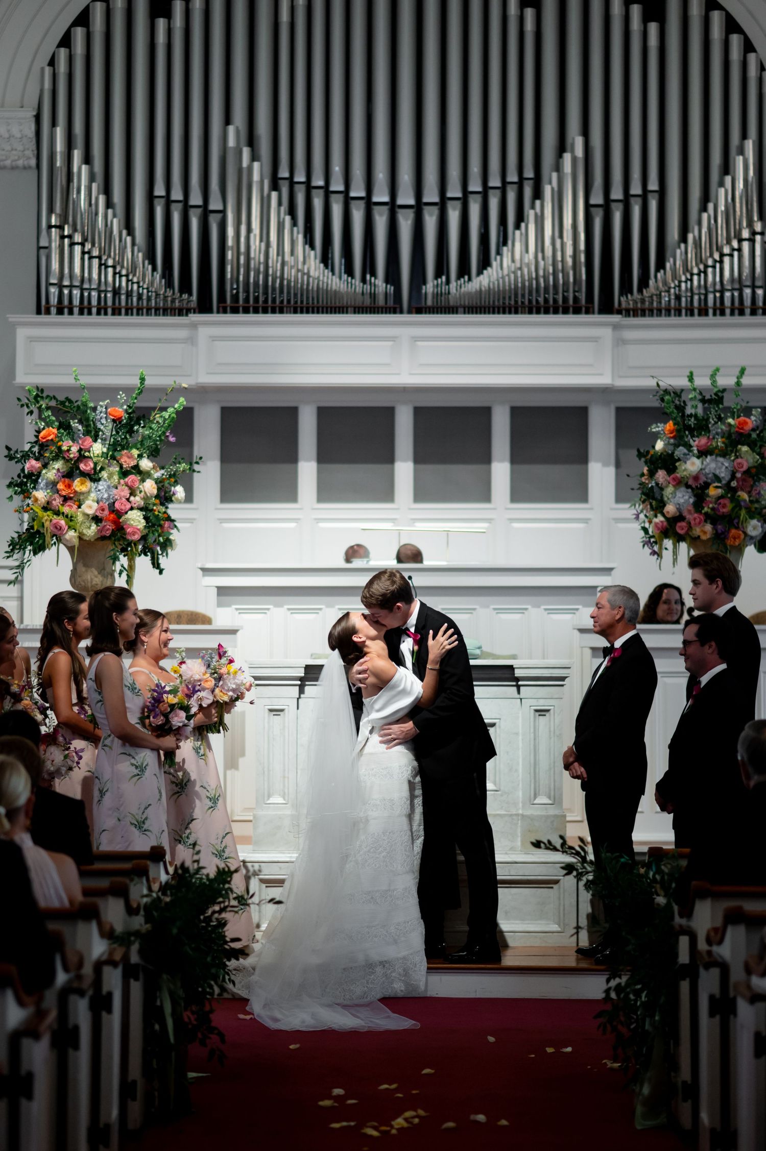 Bride and groom share first kiss at church altar with floral arrangements and pipe organ backdrop.