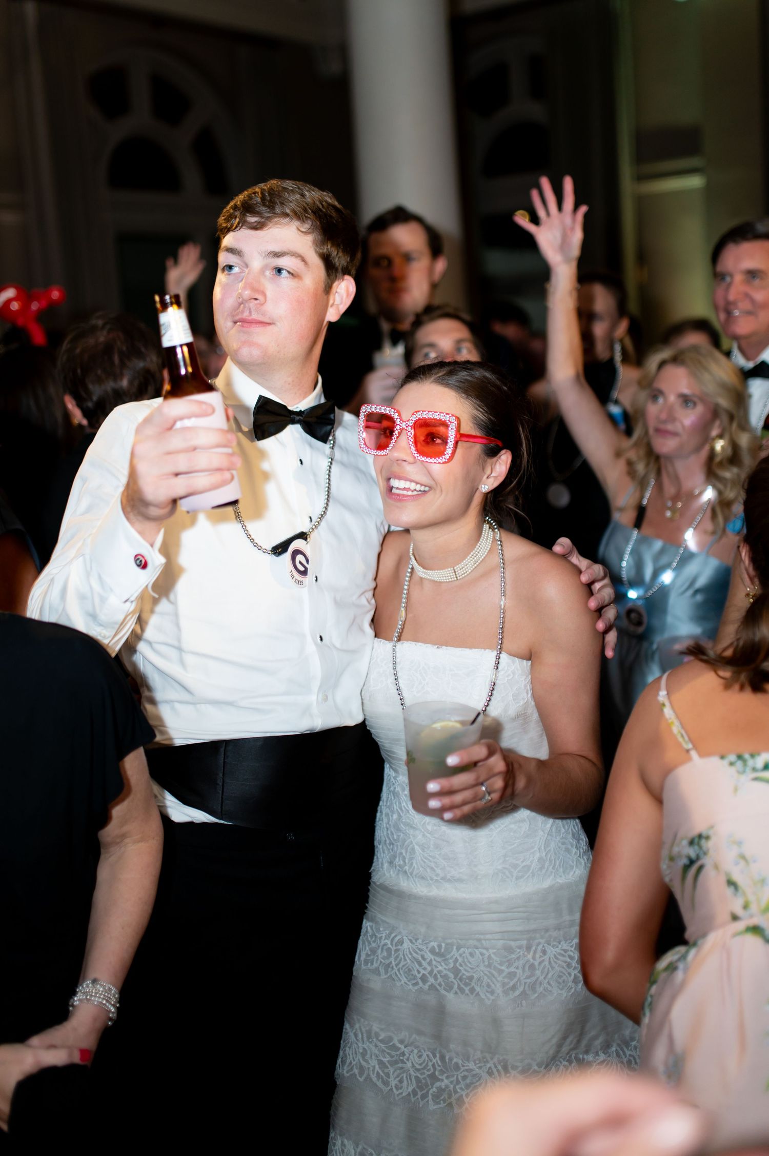 Bride wearing oversized pink sunglasses and groom dancing joyfully with guests at lively wedding reception.
