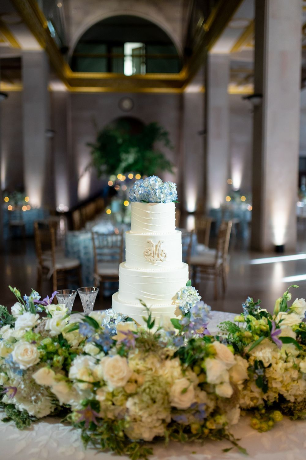 Elegant four-tier white wedding cake with gold monogram, surrounded by lush floral arrangements of blue hydrangeas and white roses.