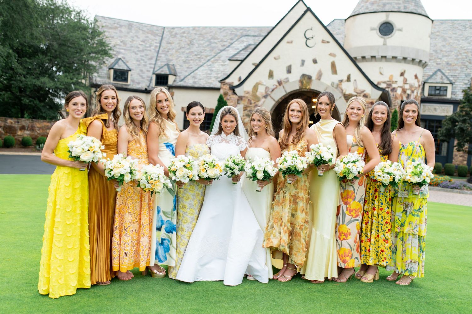 Wedding party in bright yellow dresses and white gowns stand together on grass near stone building.