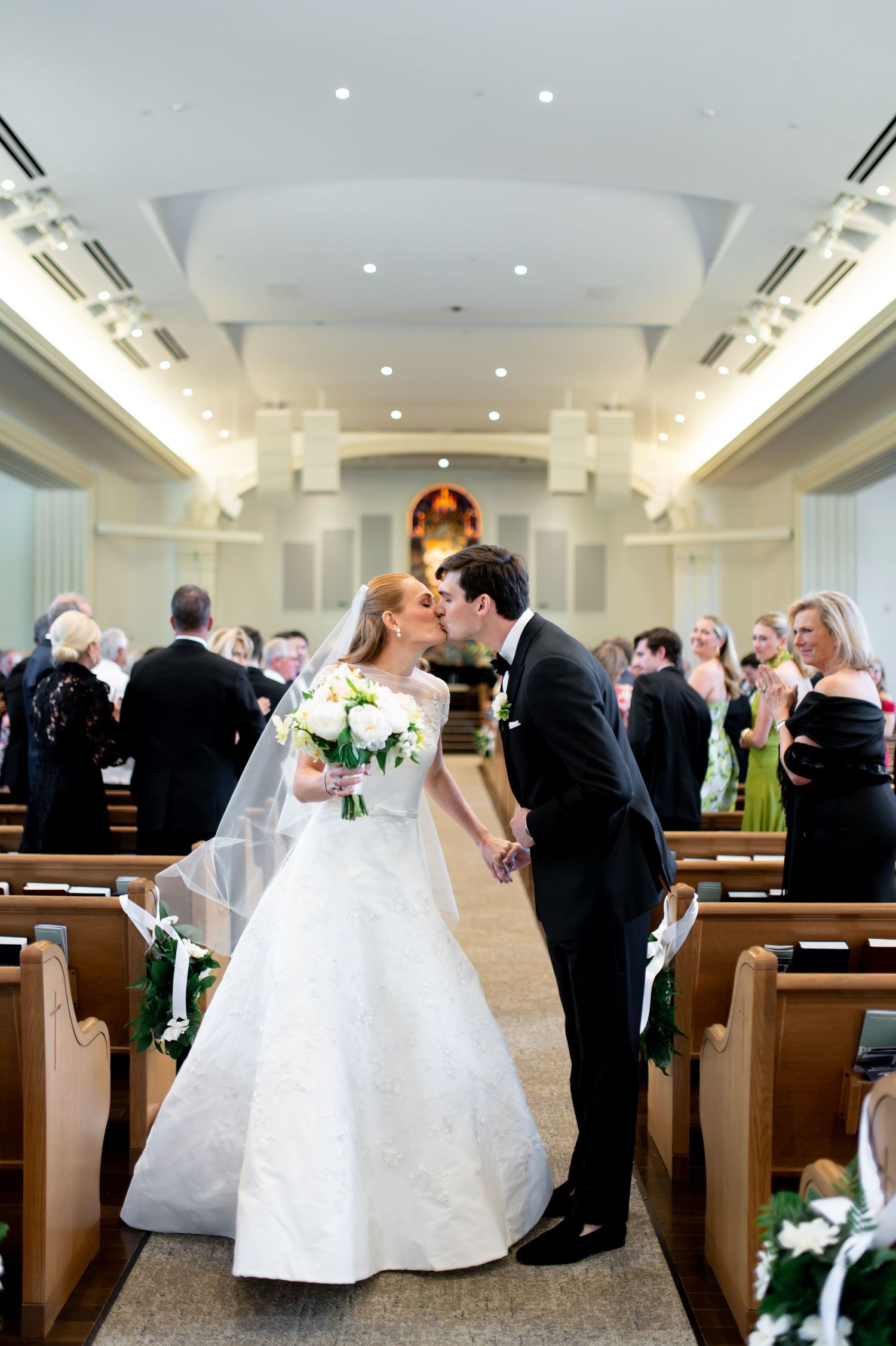 Bride and groom share first kiss walking down church aisle after wedding ceremony.