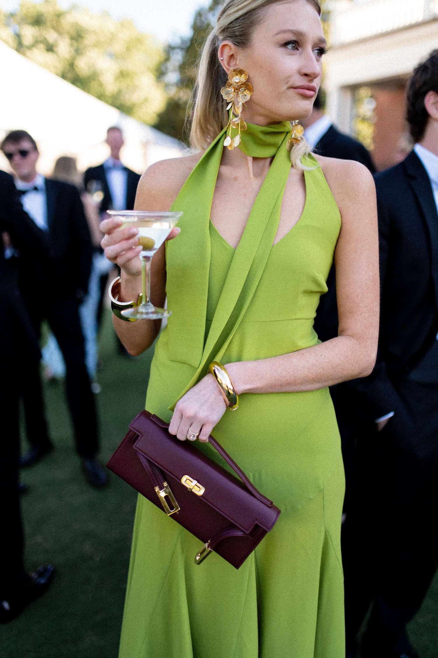Elegant wedding guest in chartreuse halter gown with gold earrings holding martini and burgundy clutch.