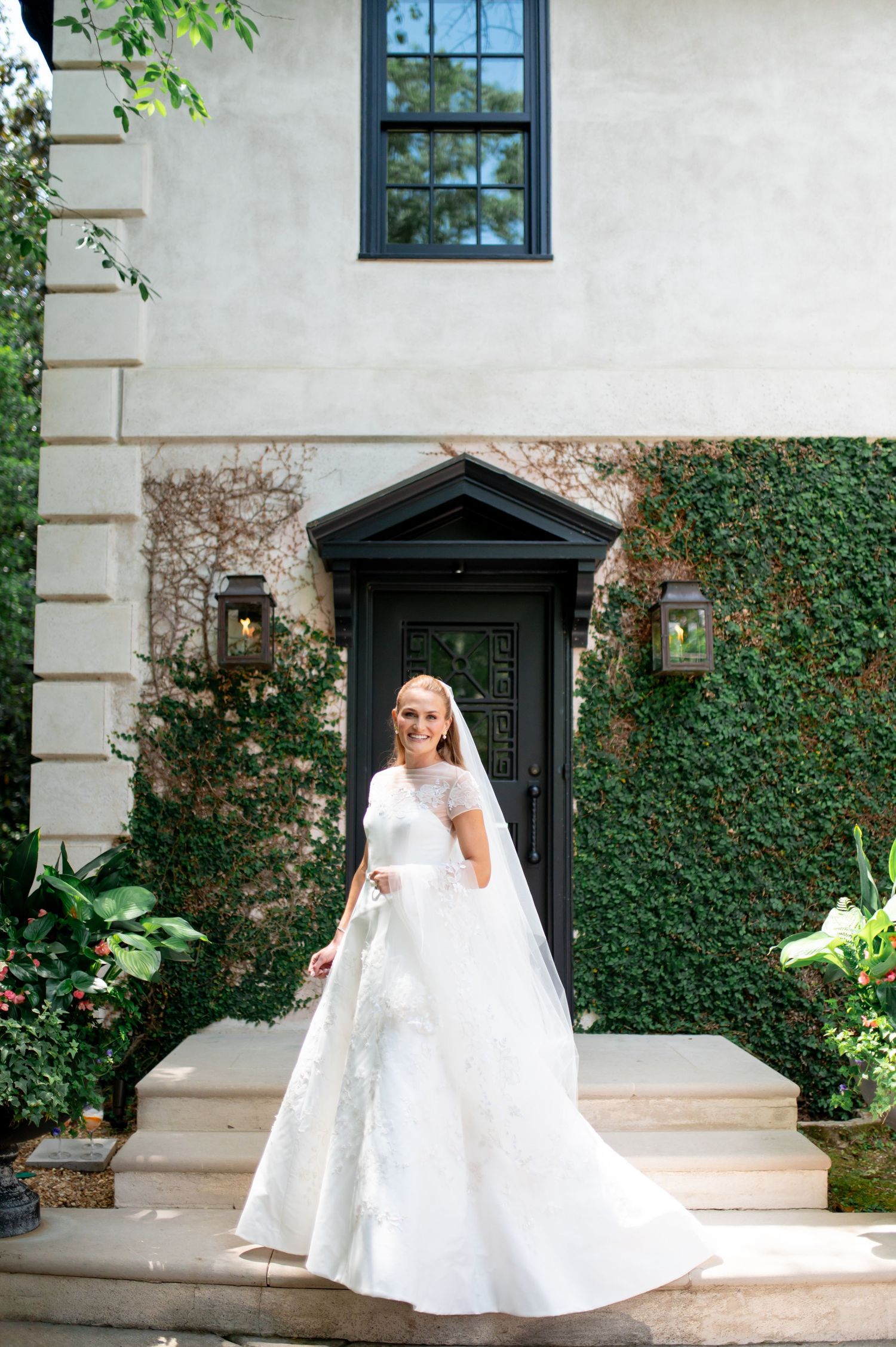 Bride in lace wedding gown and veil stands gracefully on steps of ivy-covered white estate.
