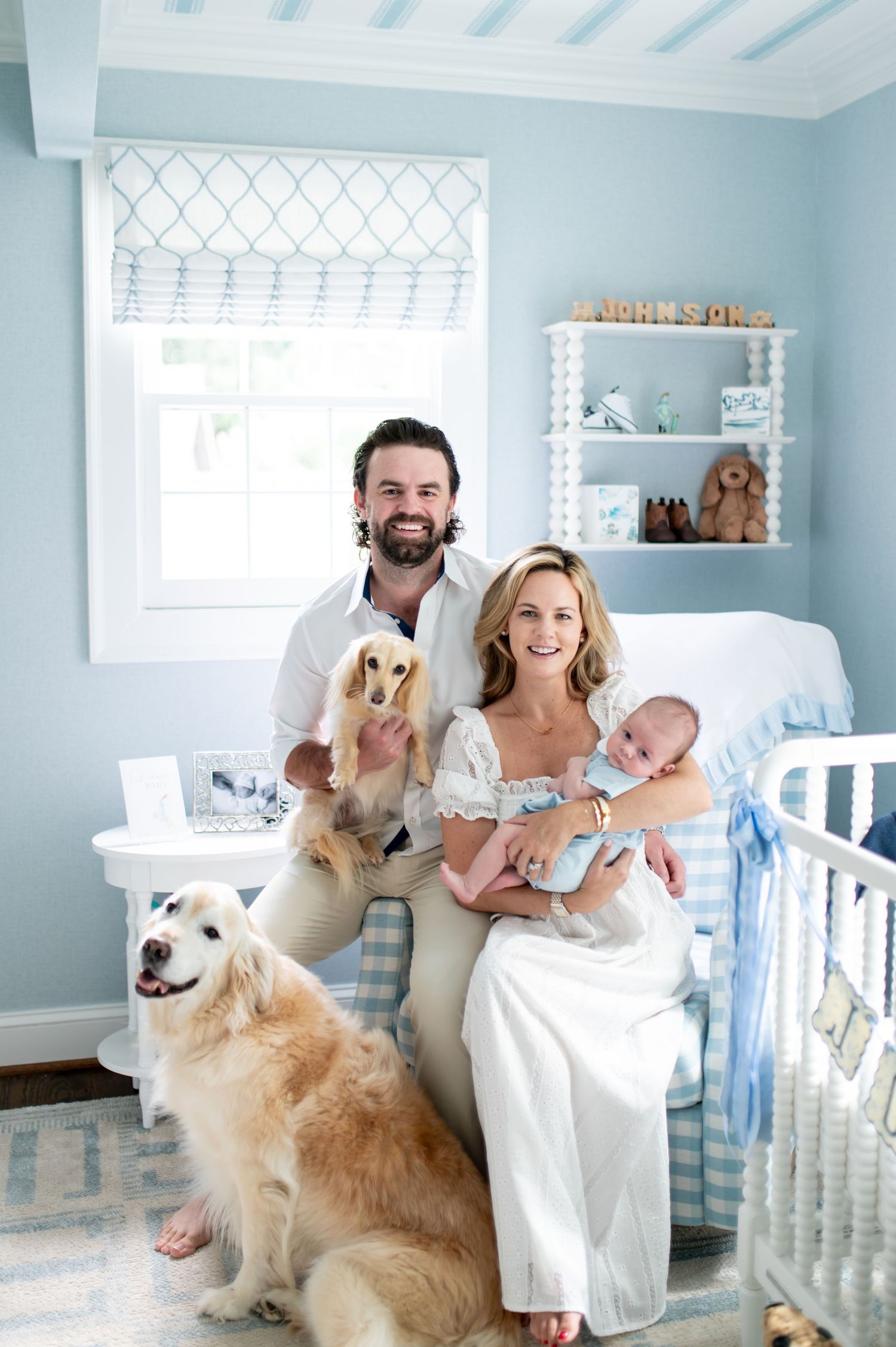 Happy family with newborn baby, two dogs, posing in a light blue nursery room with crib and shelves.