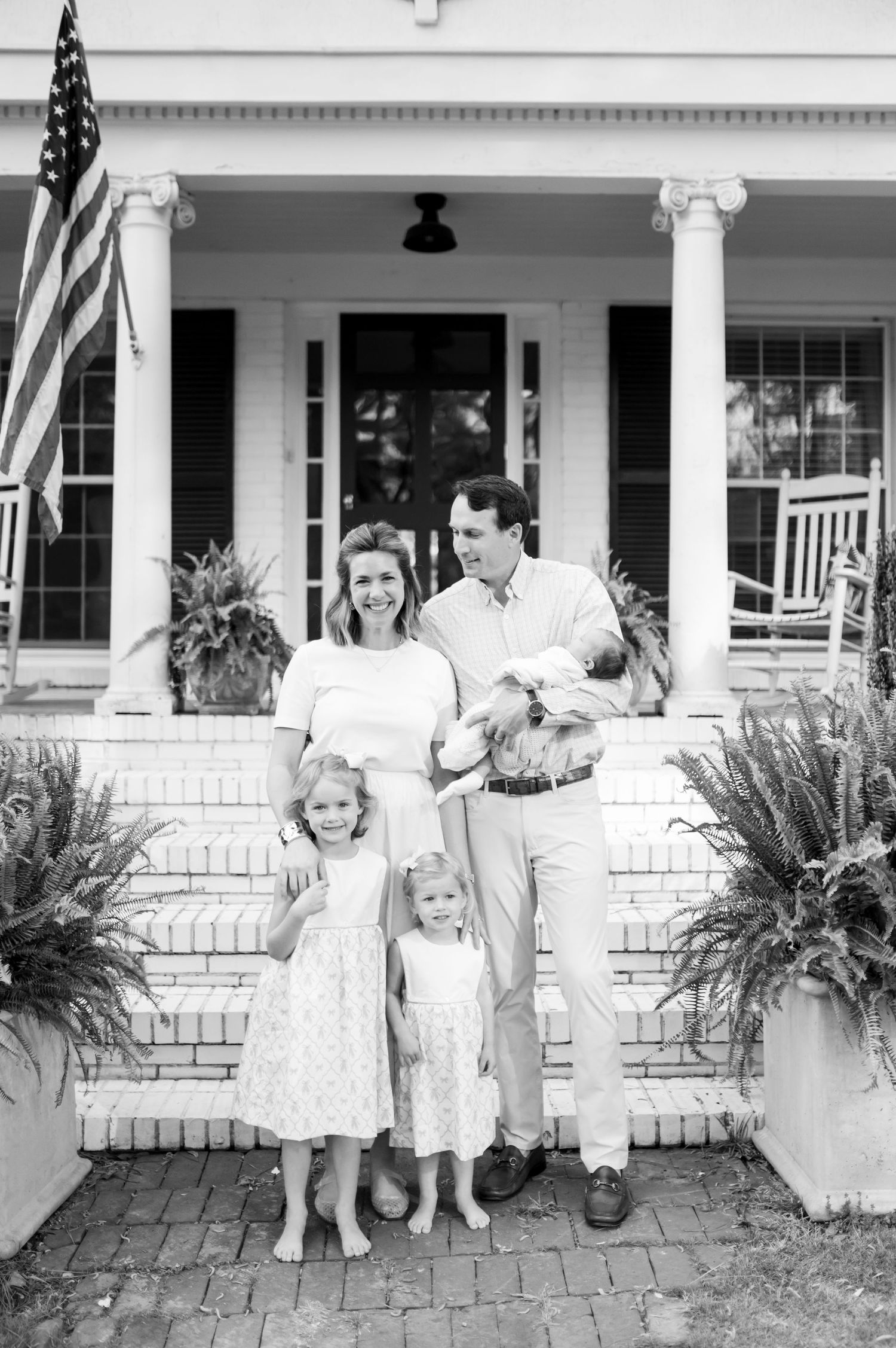 Black and white family portrait of parents with two young girls and a baby on brick porch steps of a white colonial home.