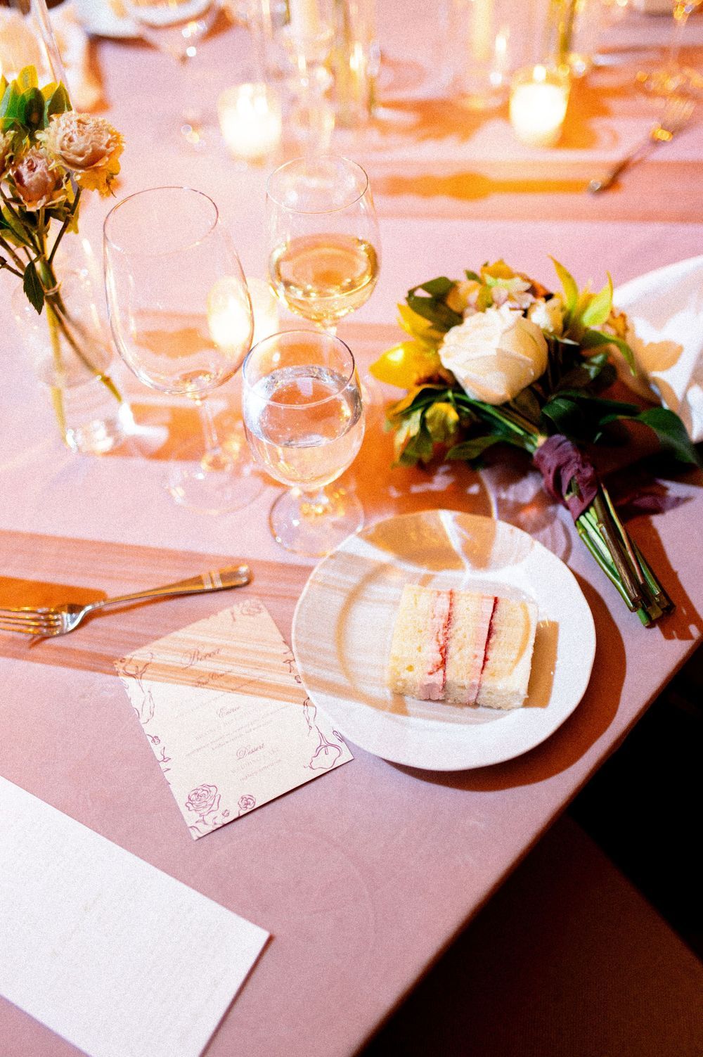 Elegant wedding reception table setting with sandwiches, wine glasses, floral bouquet, candles, and pink tablecloth.