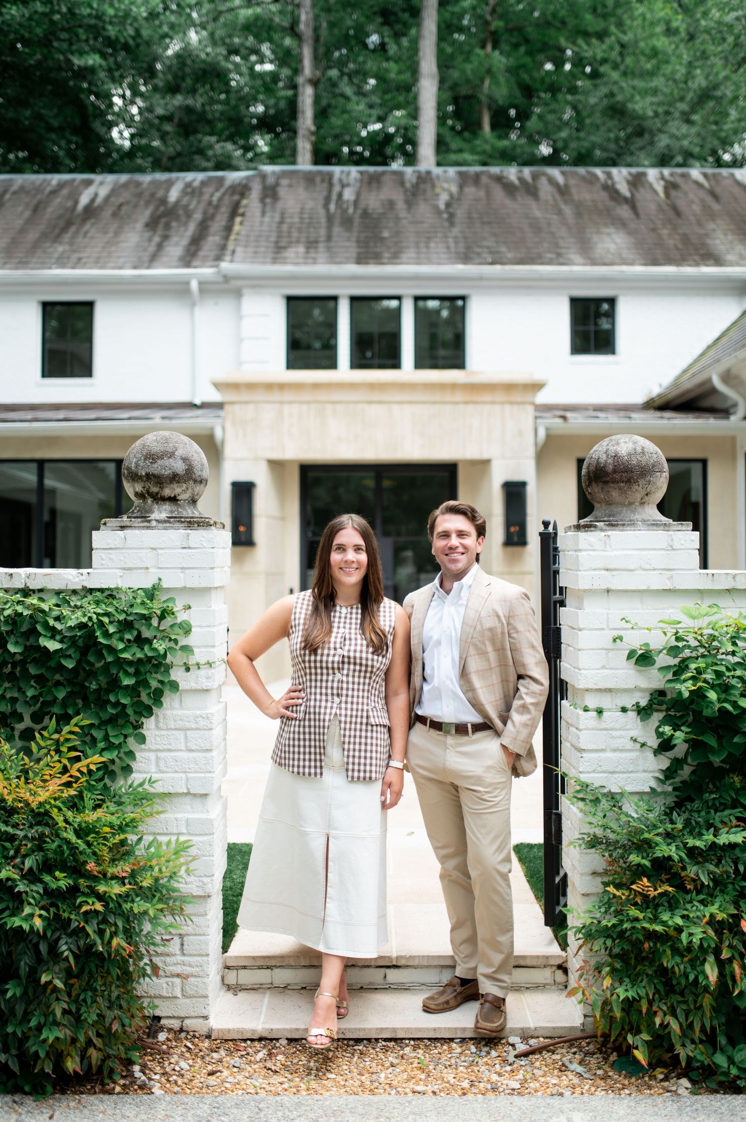 A couple stands at the entrance of a white house with stone pillars and landscaping on a summer day.