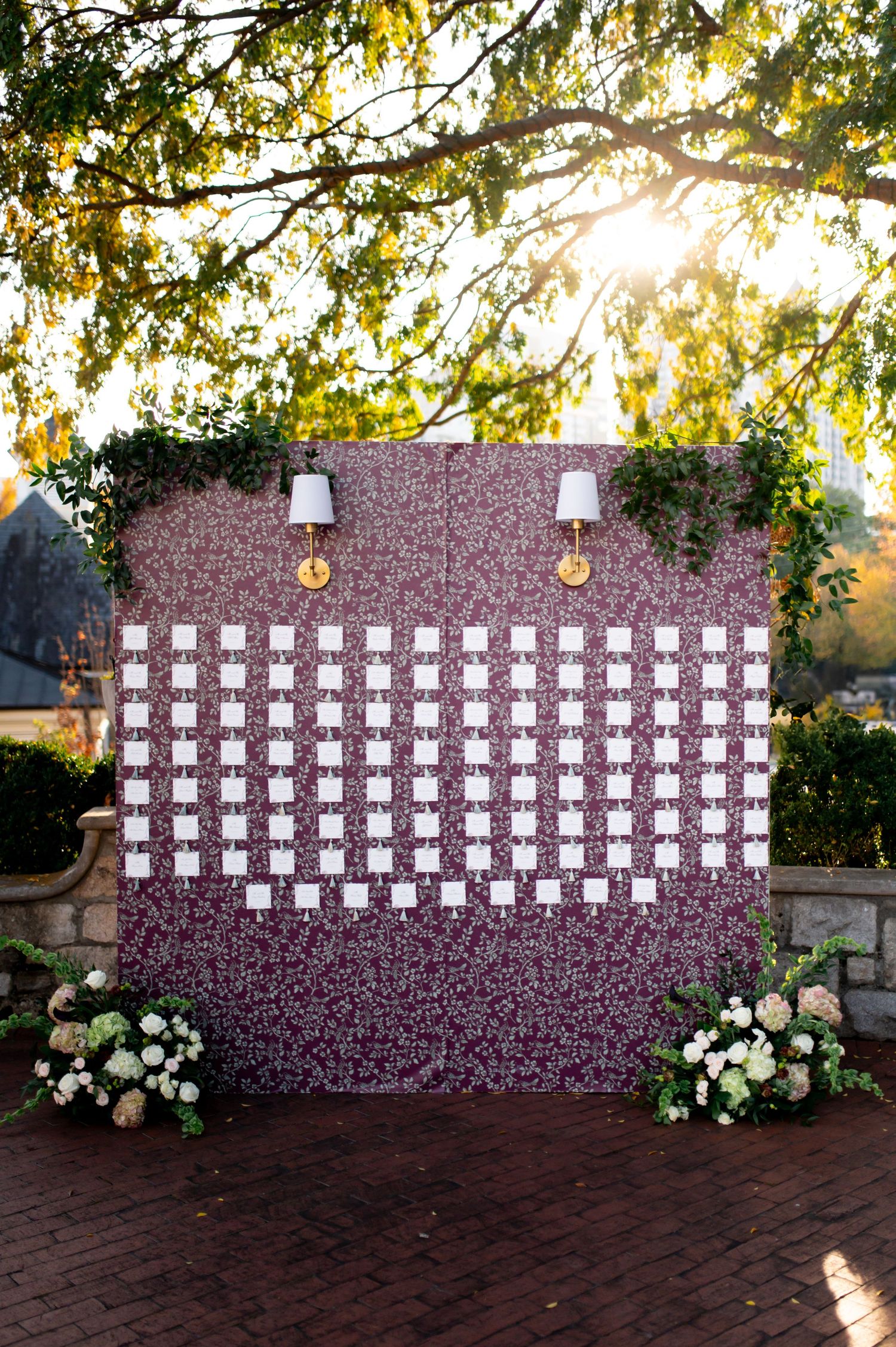 A wedding seating chart display with escort cards arranged on a rustic brick wall decorated with white floral arrangements.