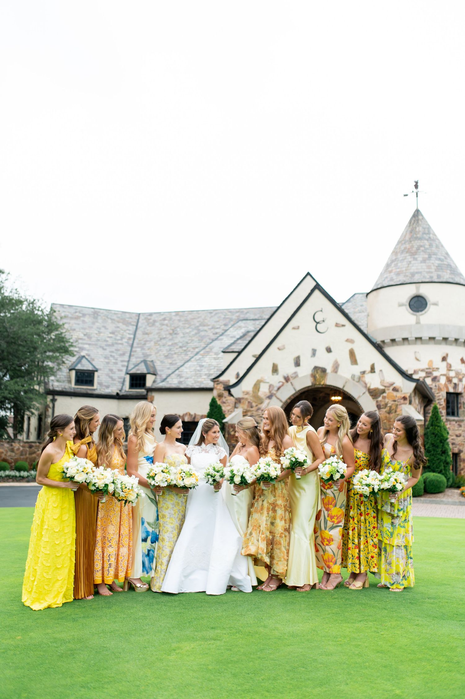 Bridesmaids in sunny yellow and floral dresses pose with bride in white on green lawn.