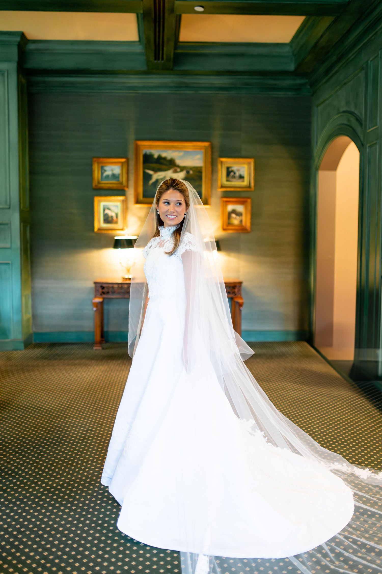 Wedding dress with flowing train displayed in an elegant room with patterned carpet and framed artwork.