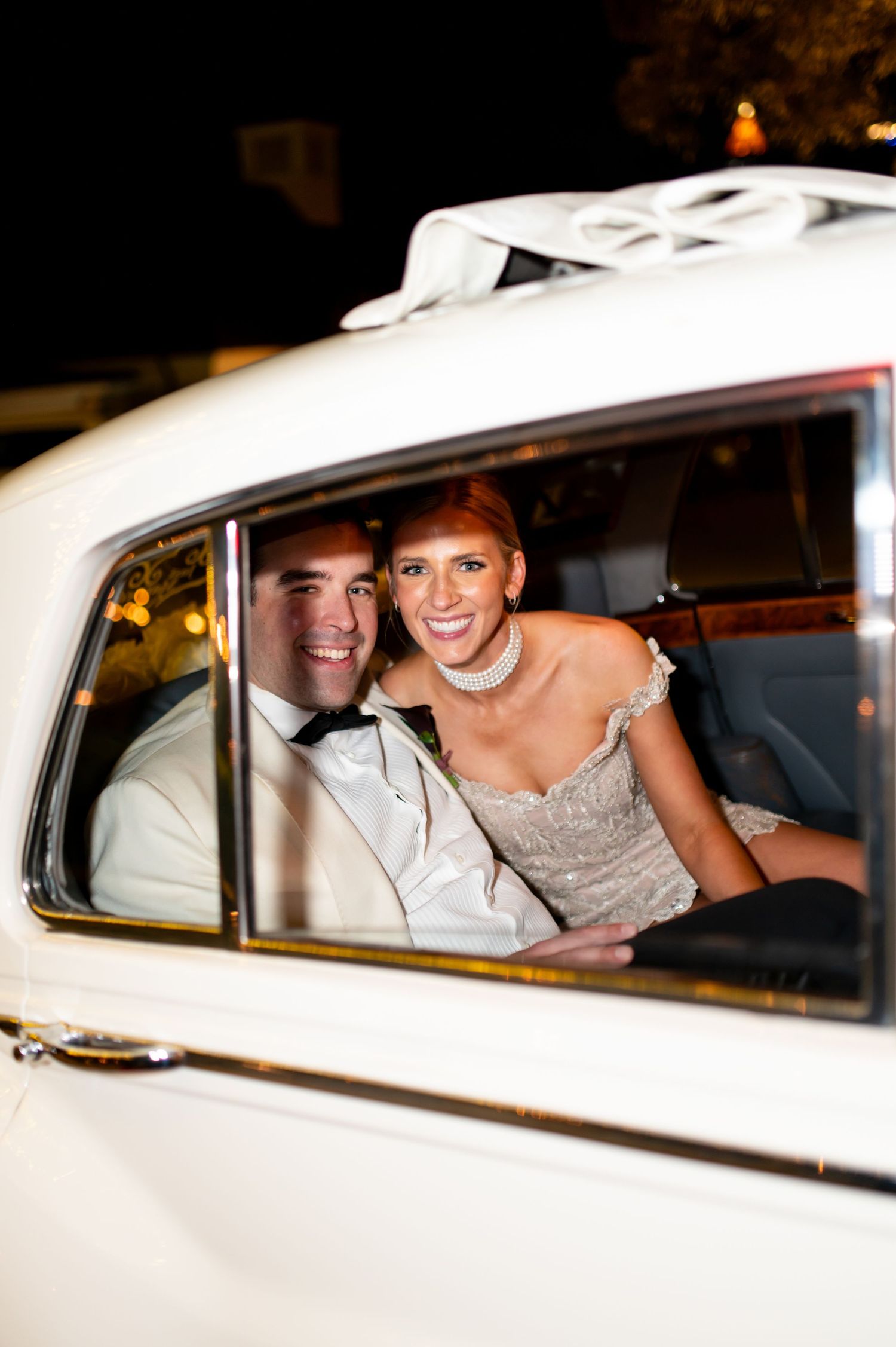 Newlyweds smiling inside classic white getaway car after reception.