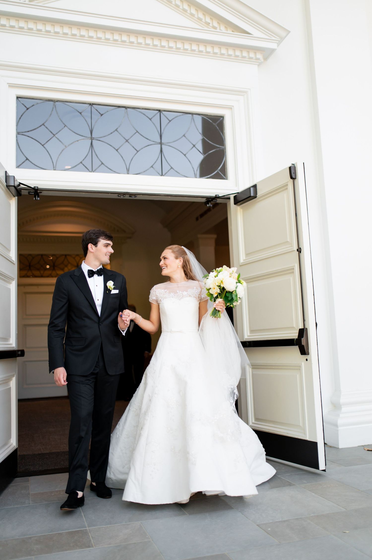 Newlyweds exit church doors, bride in lace gown holding bouquet, groom in classic black tuxedo.