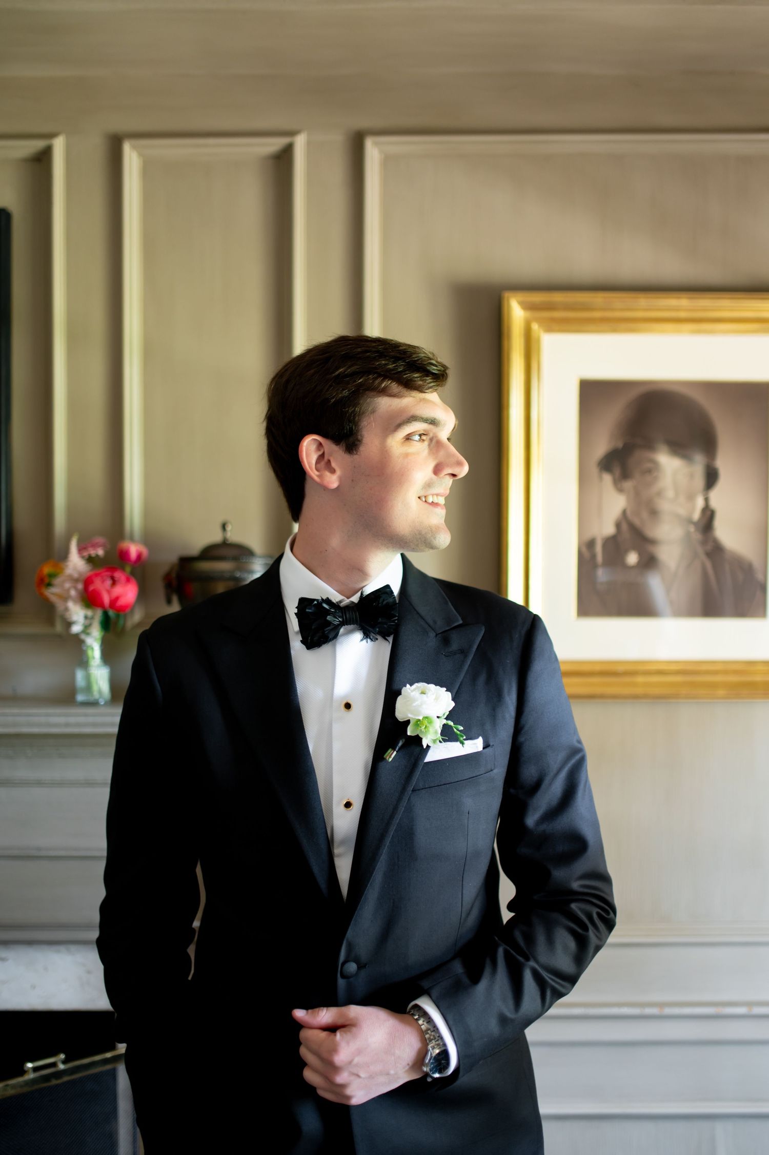 Dapper groom in black tuxedo with bow tie and white boutonniere stands before framed portrait.