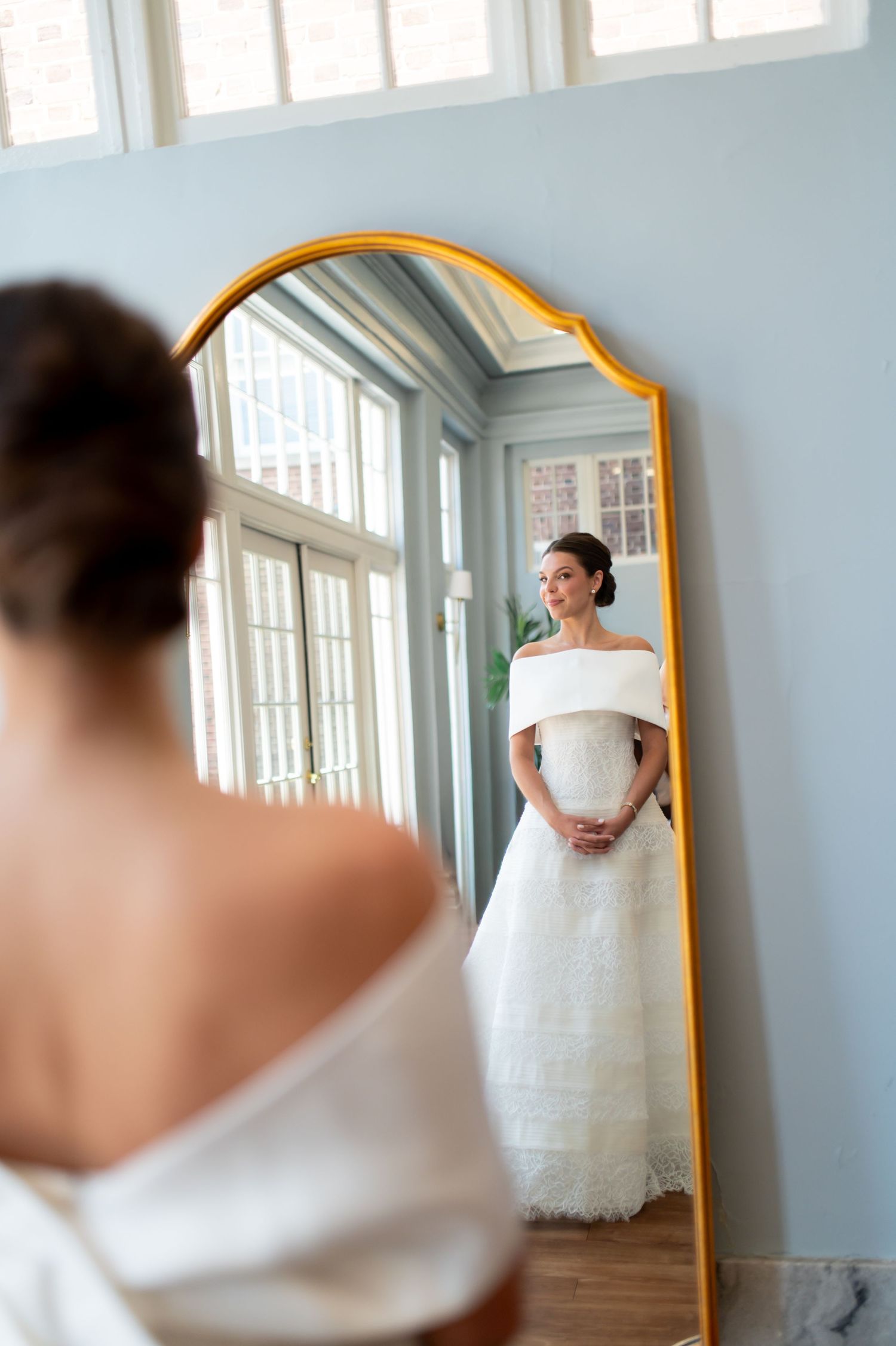 Bride in off-shoulder white wedding gown admiring her reflection in a gold-framed floor mirror.