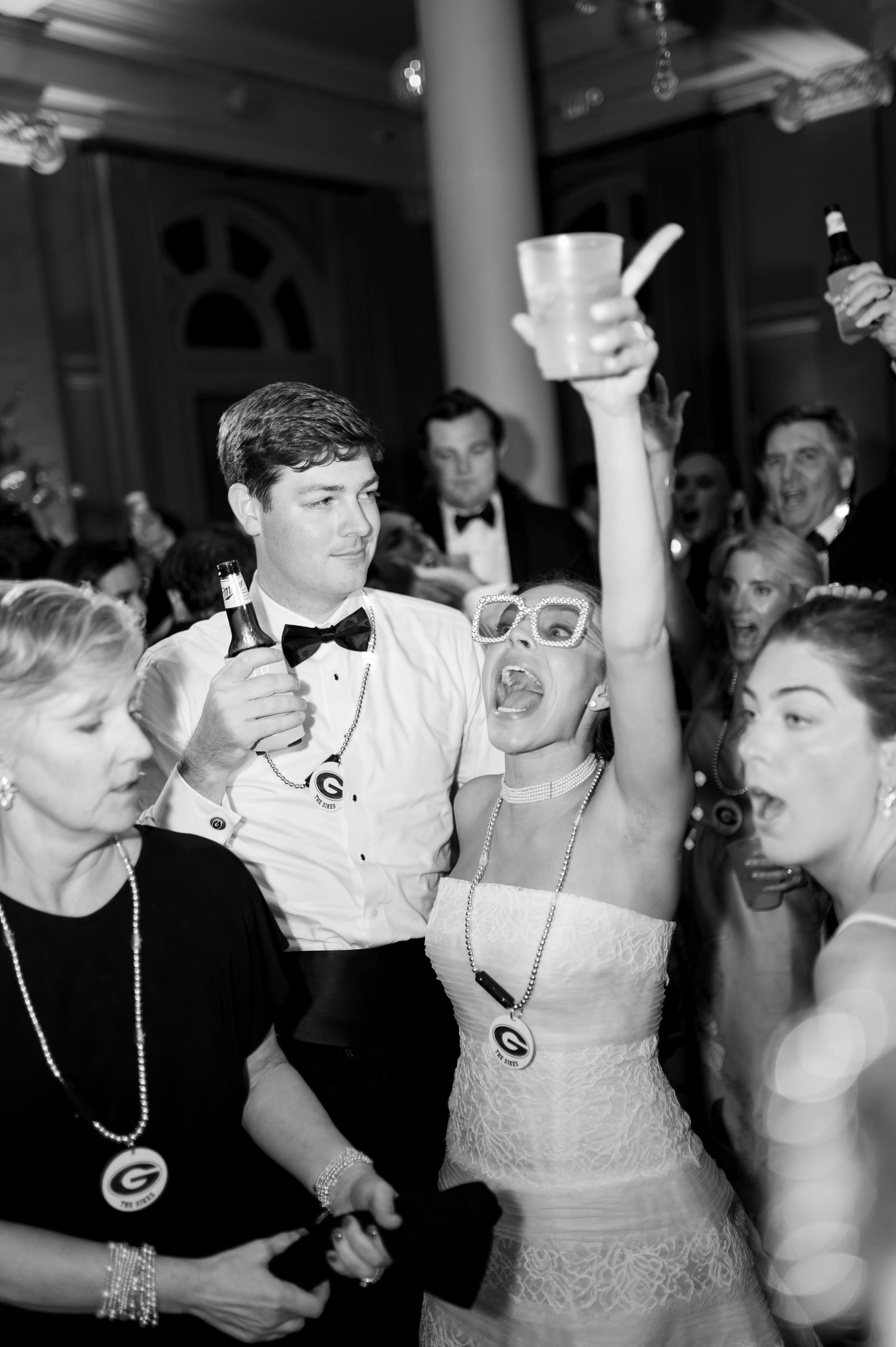 Black and white photo of bride raising cup celebrating on dance floor with guests at reception.
