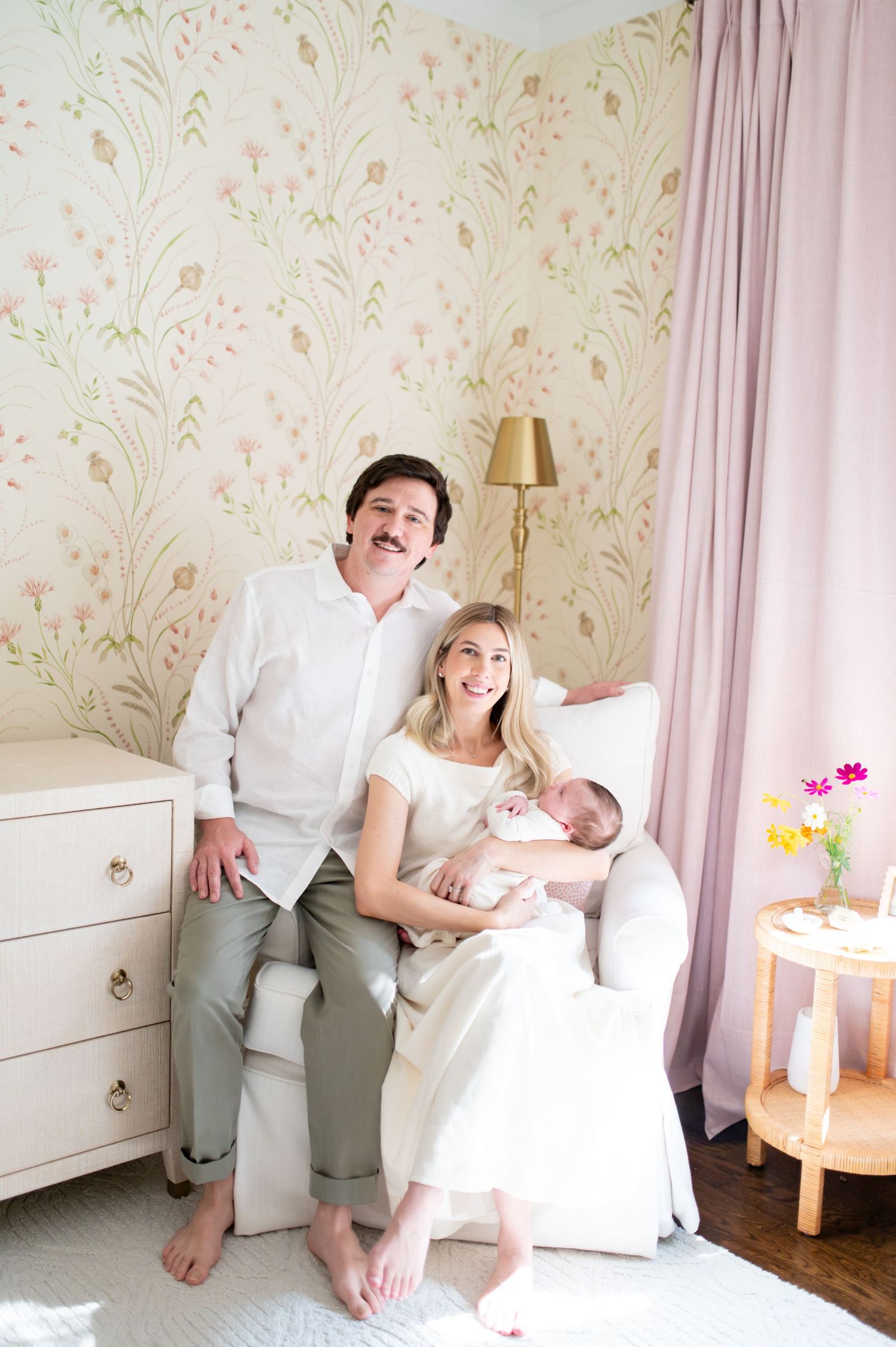 New parents smiling with their newborn baby in a beautifully decorated nursery with floral wallpaper and pink curtains.
