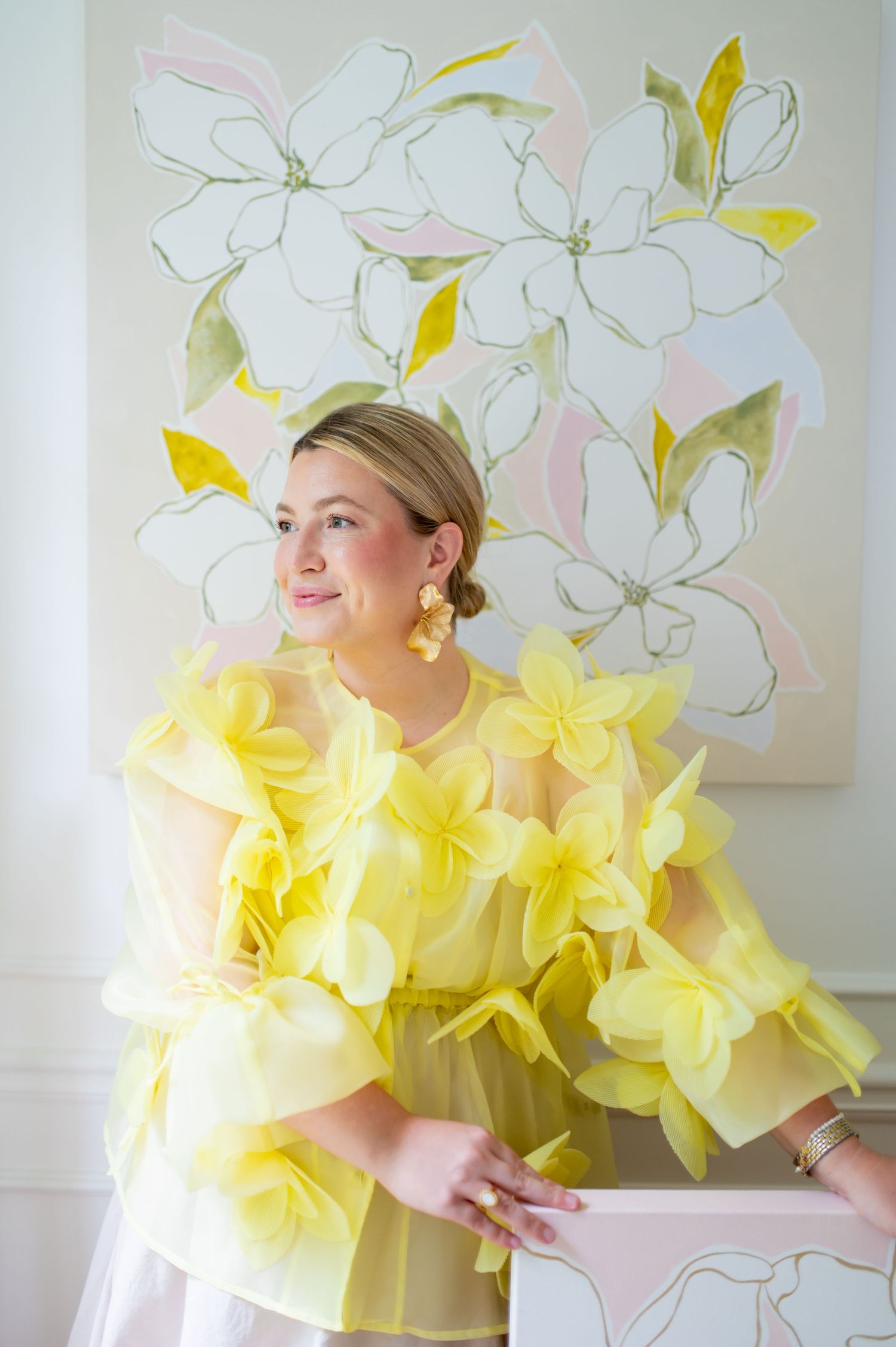 Woman in yellow floral organza dress poses before large magnolia painting, wearing gold earrings and bracelet.