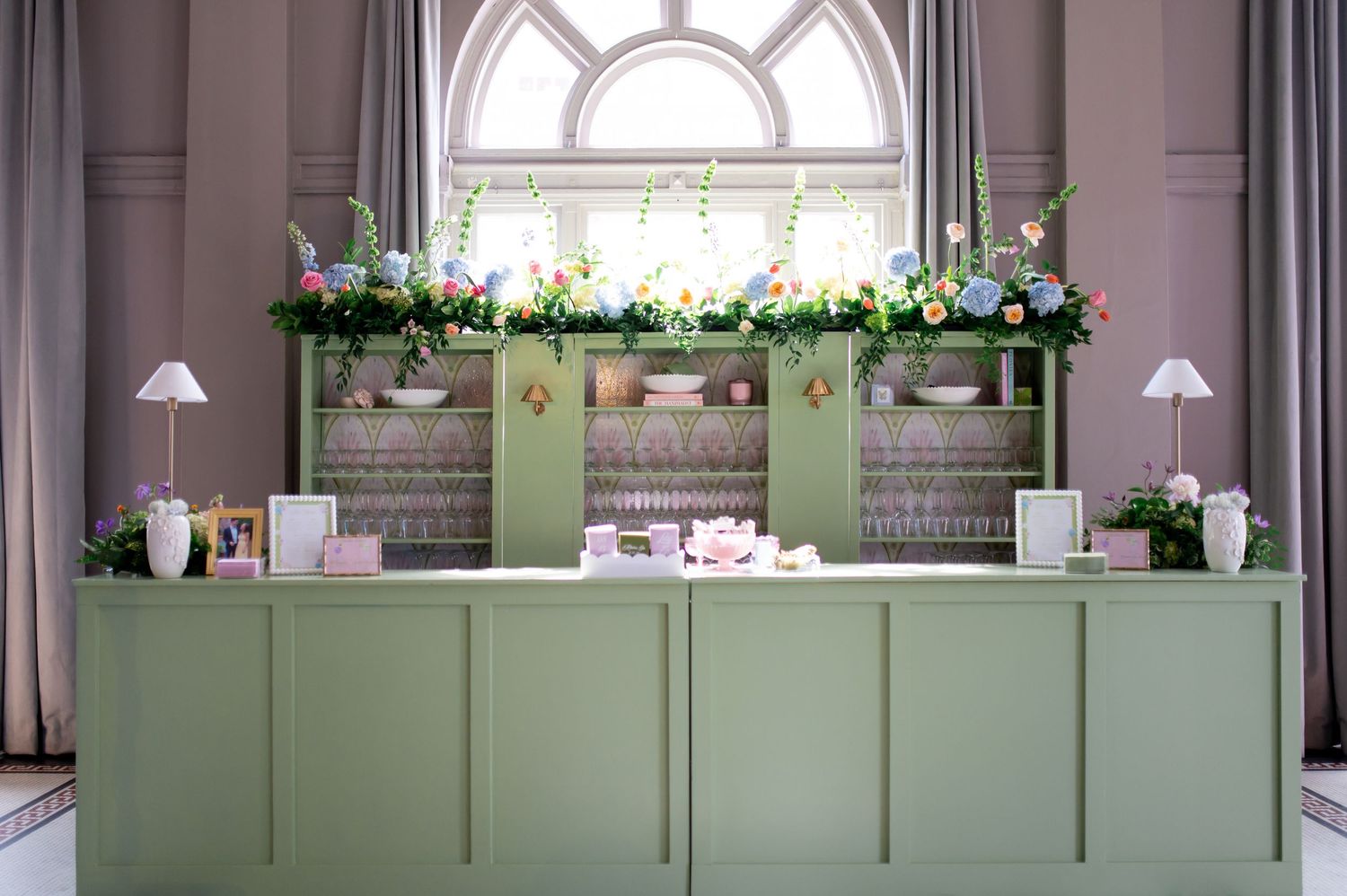 Sage green reception bar decorated with vibrant floral arrangements displayed beneath a grand arched window.