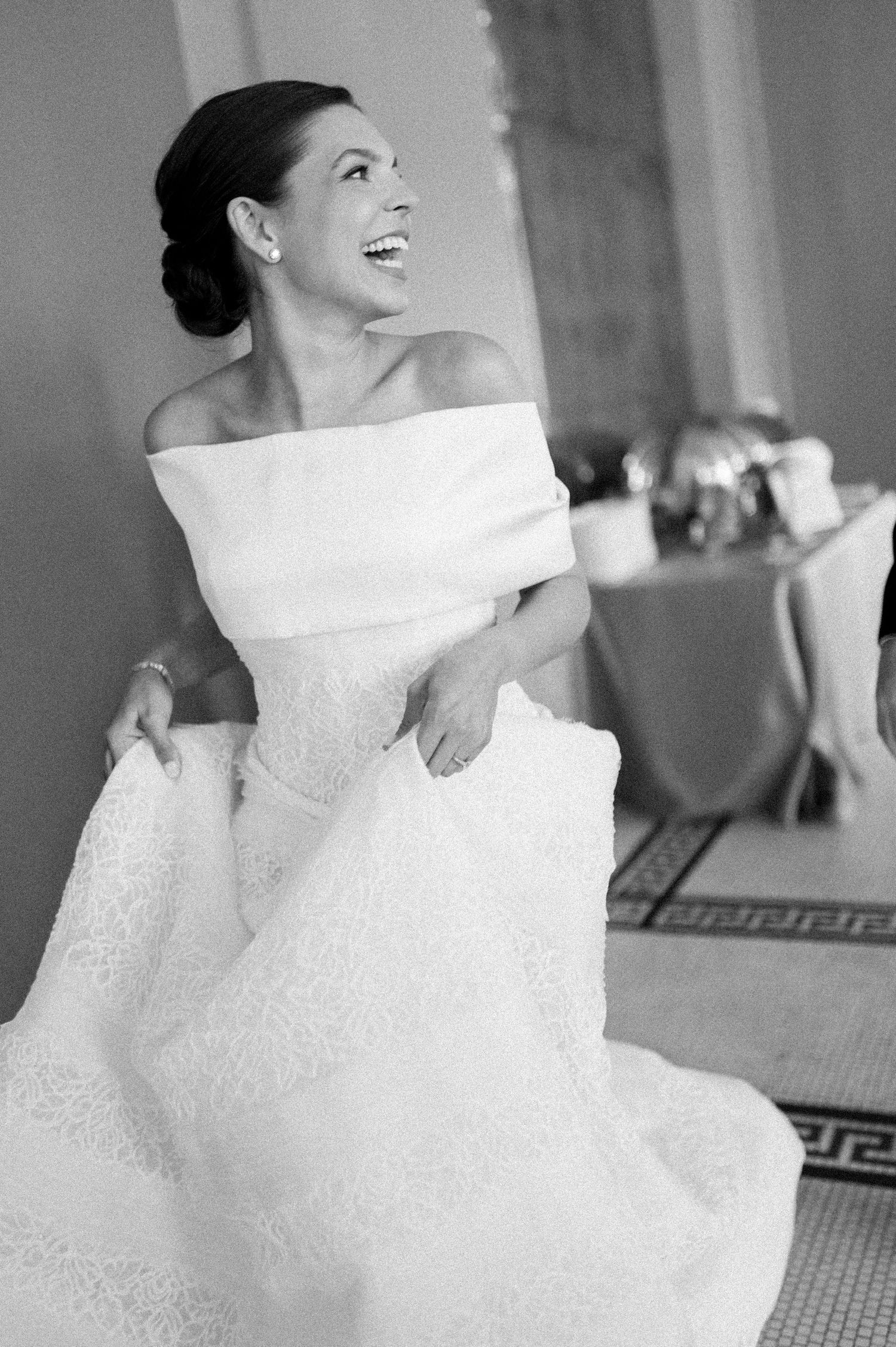 Bride in off-shoulder white lace ball gown laughing joyfully in black and white wedding photo.