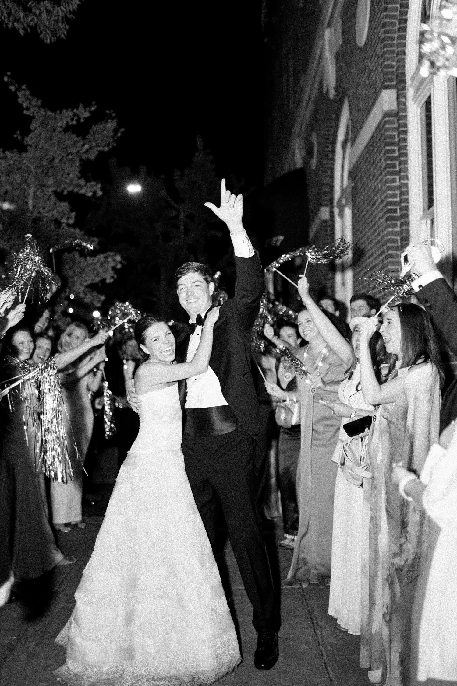 Bride and groom exit their wedding reception in black and white photo as guests cheer them on.