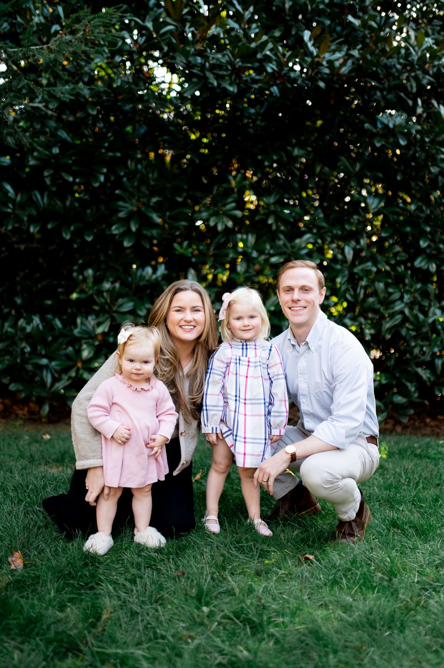 A smiling family of four poses outdoors on green grass with lush shrubs, featuring two young girls in pink and plaid dresses.
