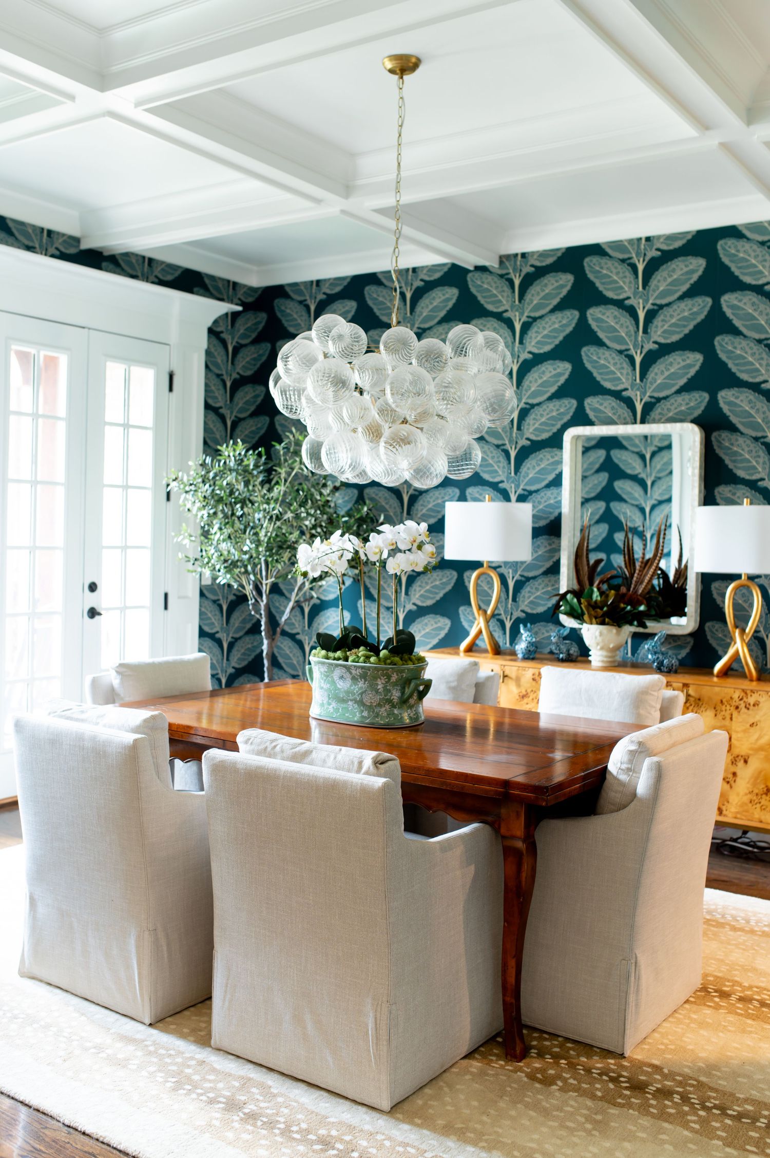 Modern dining room features teal leaf wallpaper, white upholstered chairs, and a wood table under bubble glass chandelier.