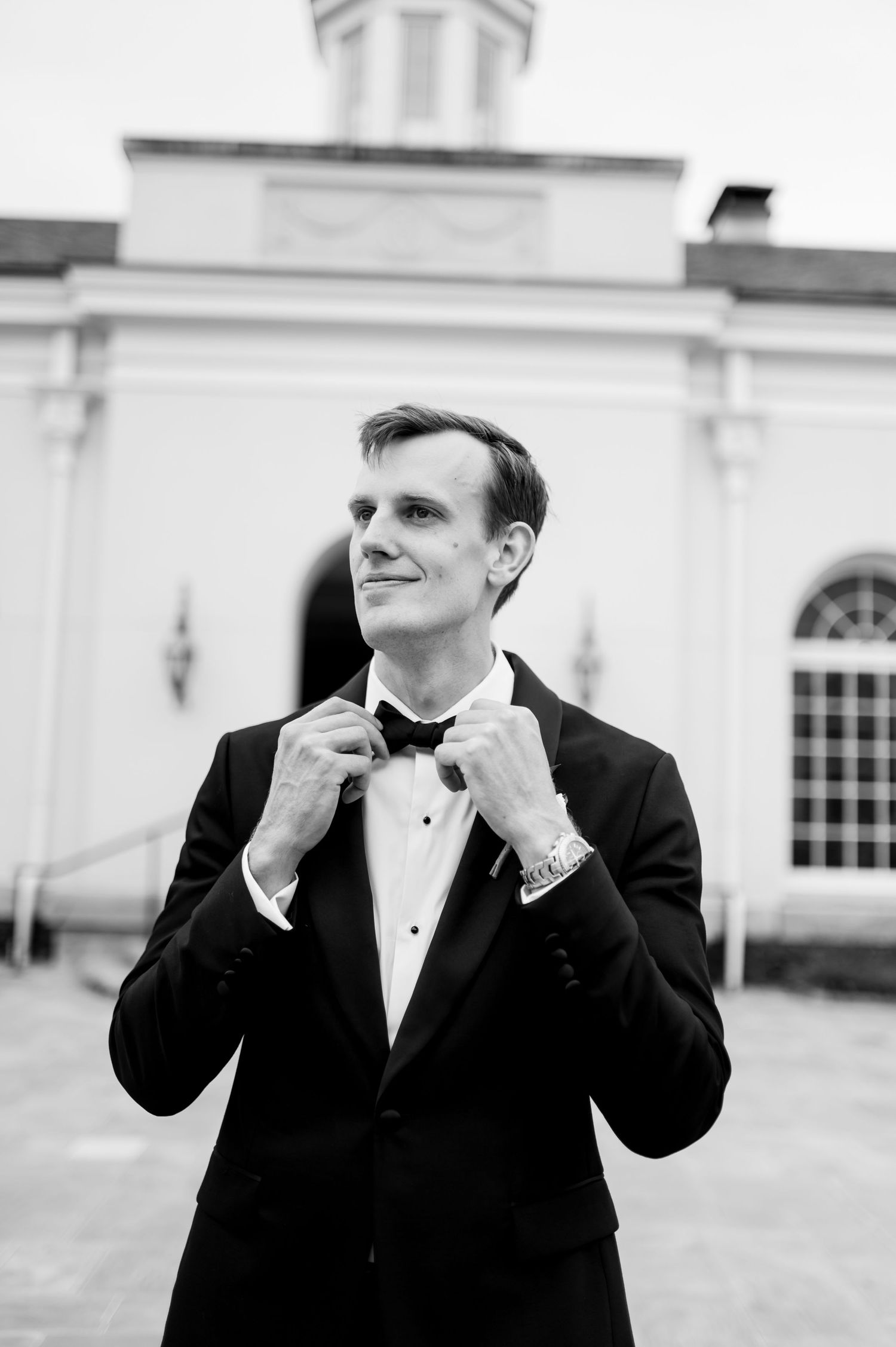 Groom in classic black tuxedo adjusts bowtie in black and white portrait.