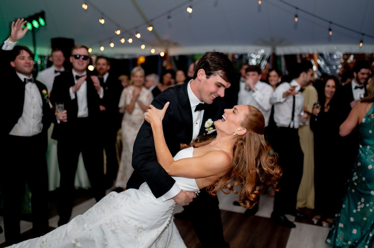 Groom dips bride on dance floor surrounded by cheering guests at tented wedding reception.