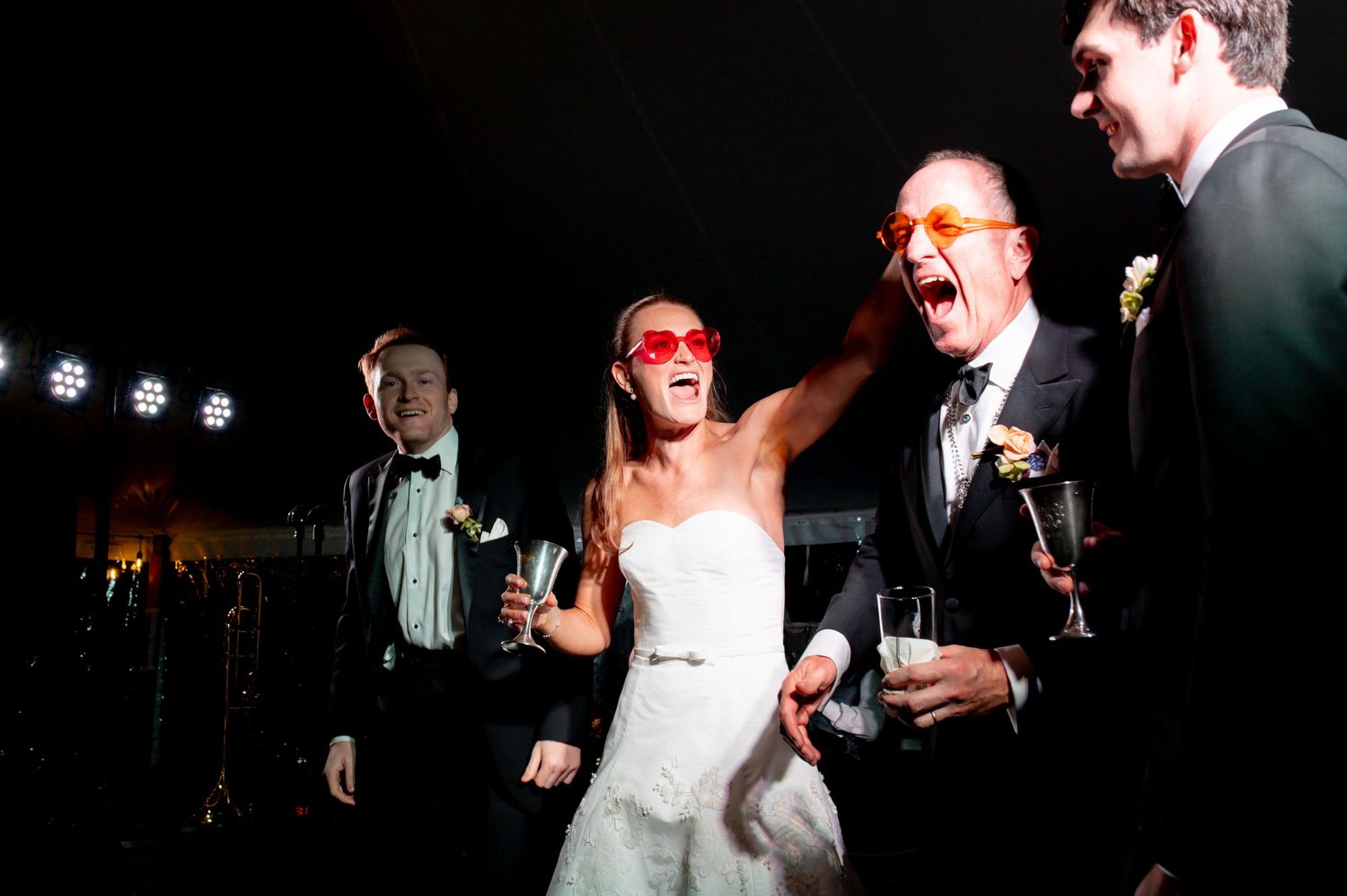 Bride and guests laugh and celebrate wearing red sunglasses during energetic wedding reception party.