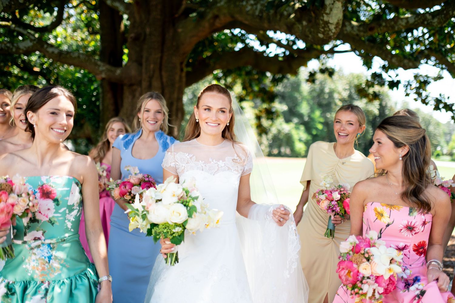 Bride in lace gown leads bridesmaids in colorful floral dresses carrying vibrant bouquets outdoors.