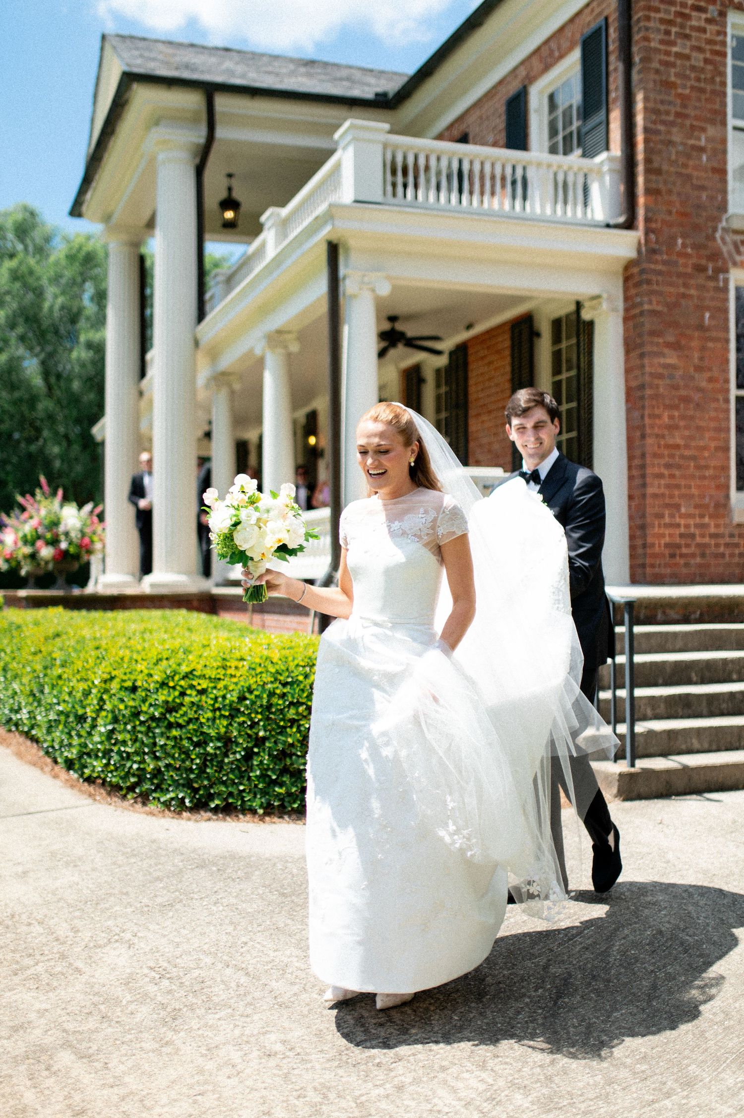 Joyful bride in white gown carries bouquet as groom in tuxedo follows outside brick mansion.