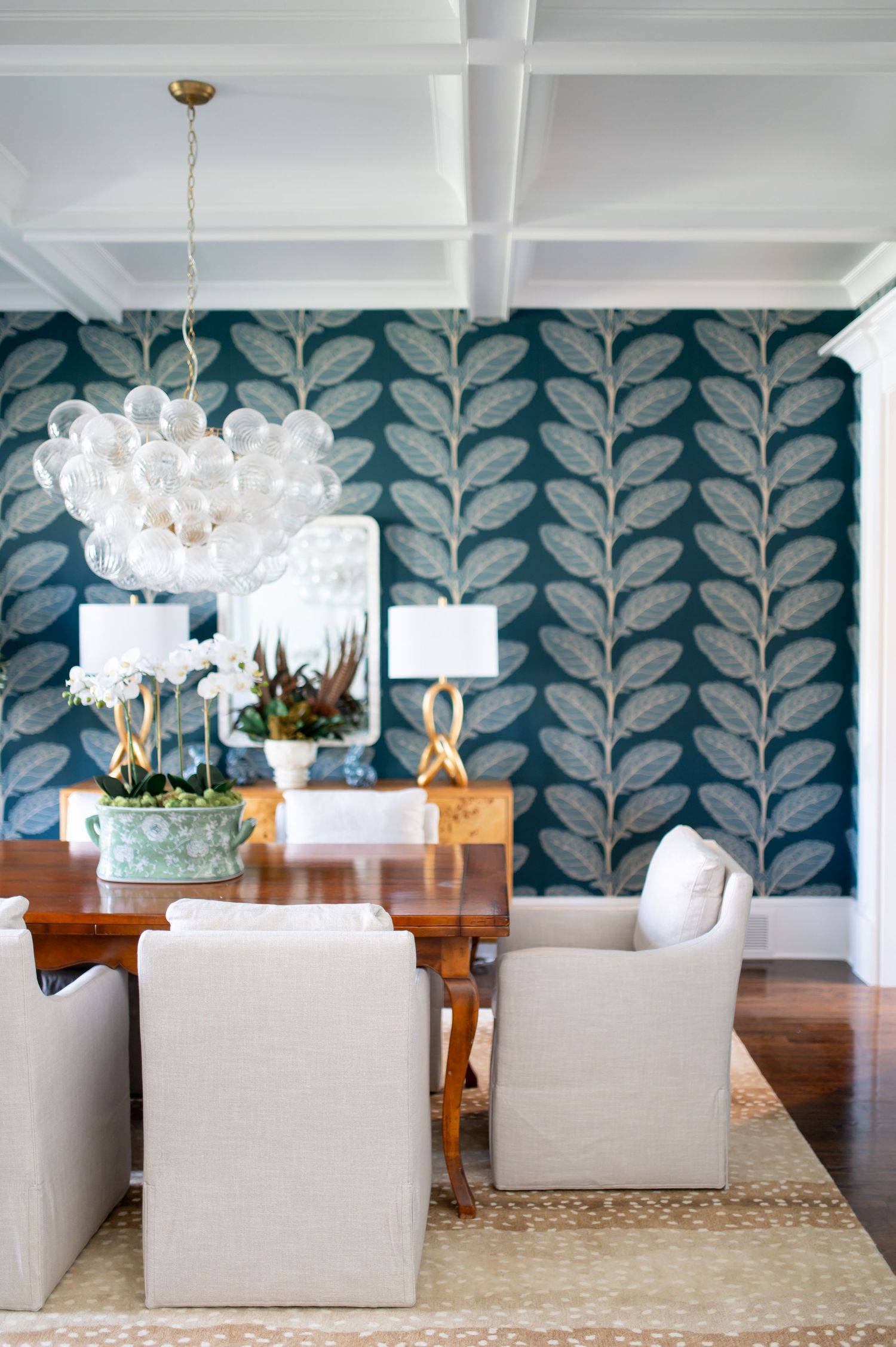 Modern dining room features blue leaf wallpaper, cream upholstered chairs, and elegant glass chandeliers above a wooden table.