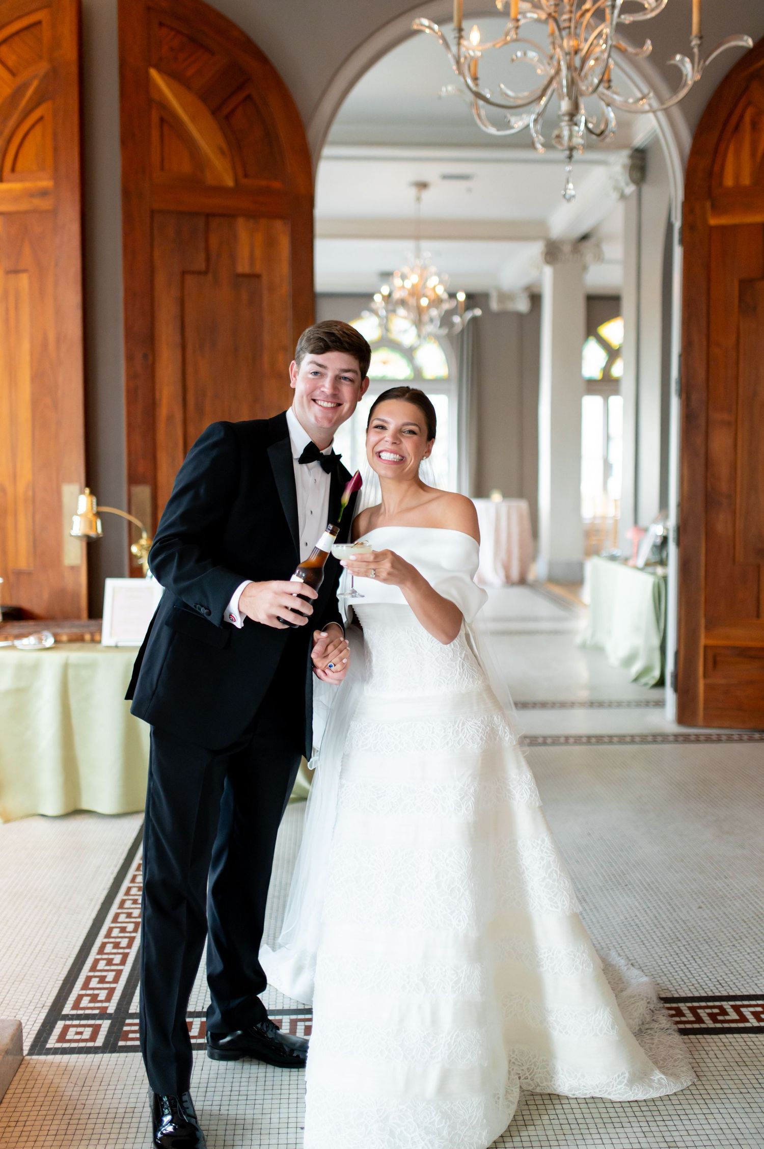 Smiling bride and groom celebrate with champagne bottle in elegant venue with chandeliers and arched doorways.