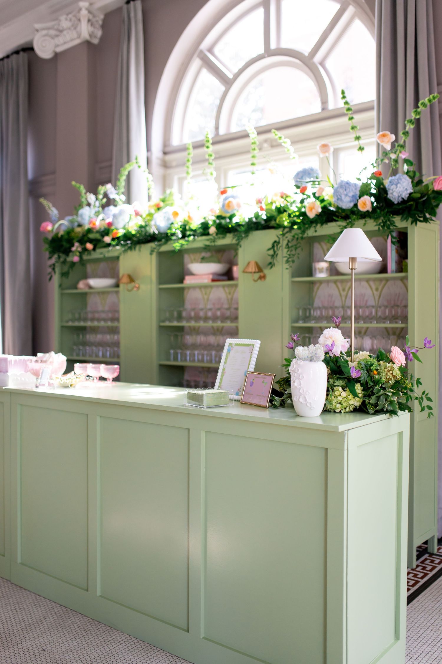 Sage green wedding bar with pink floral arrangements and lamp accents beneath an elegant arched window.