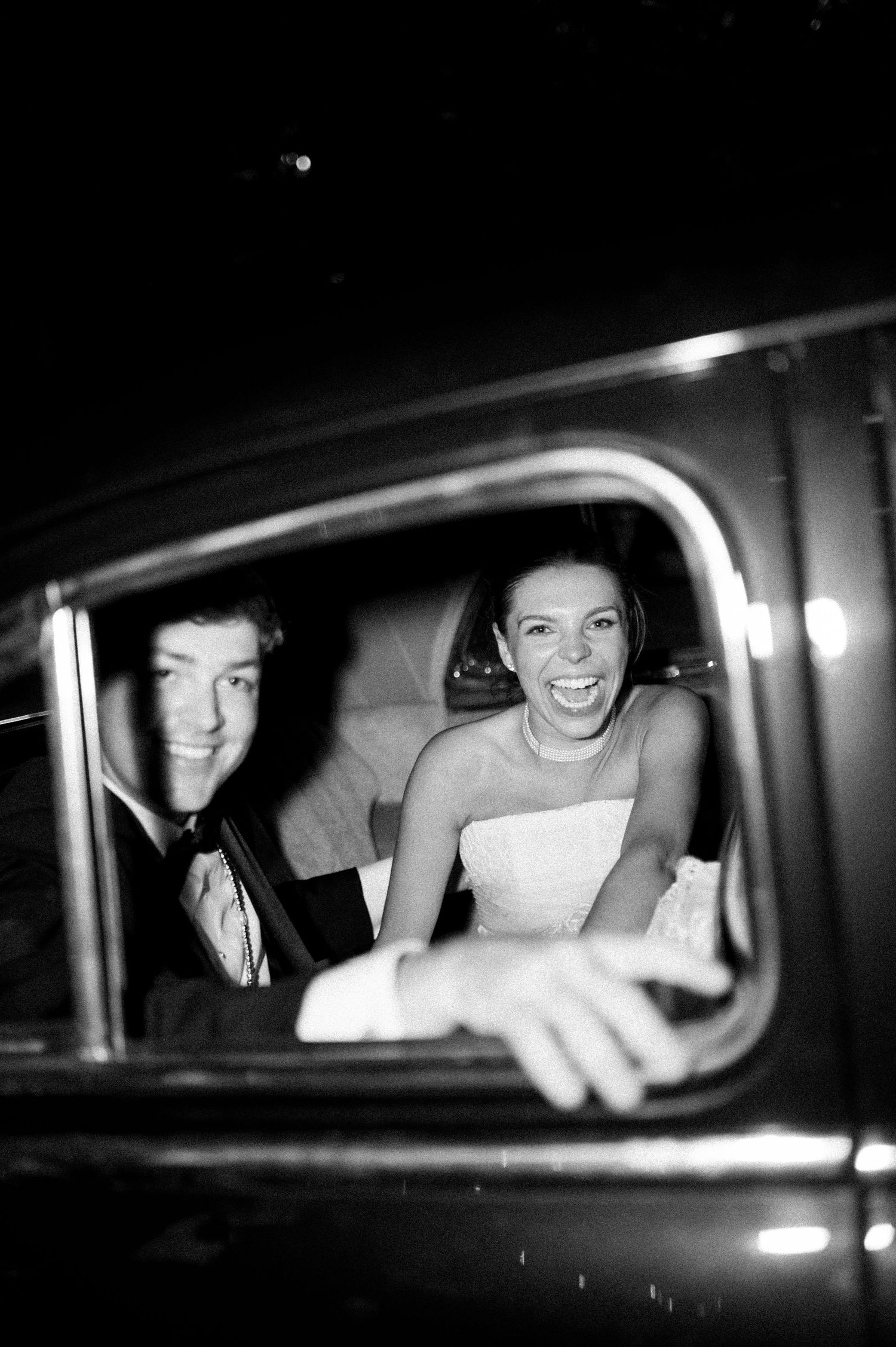 Black and white photo of laughing bride and groom sitting together in vintage car after wedding.