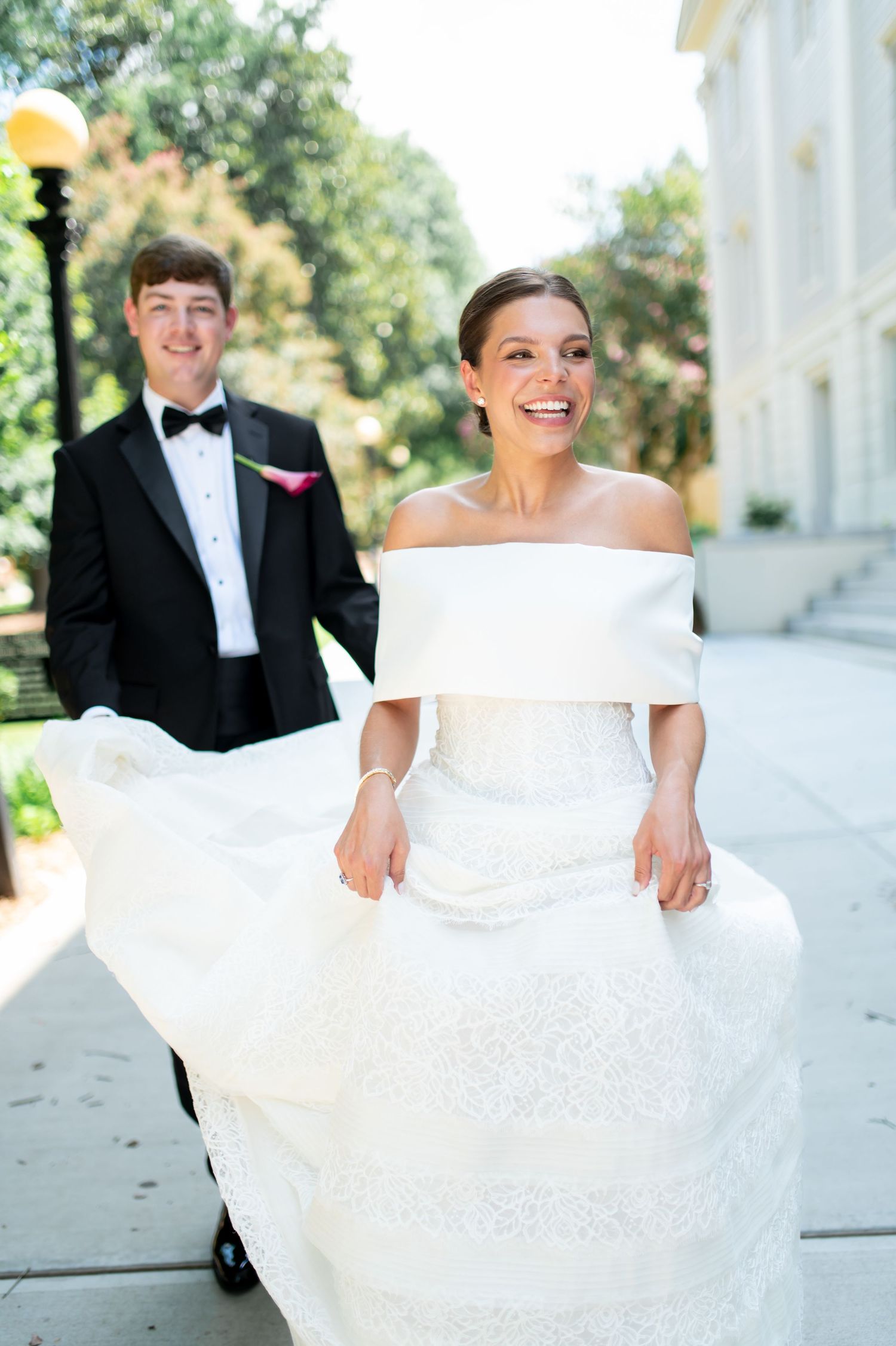 Joyful bride in off-shoulder lace gown walks with smiling groom in black tuxedo outdoors.