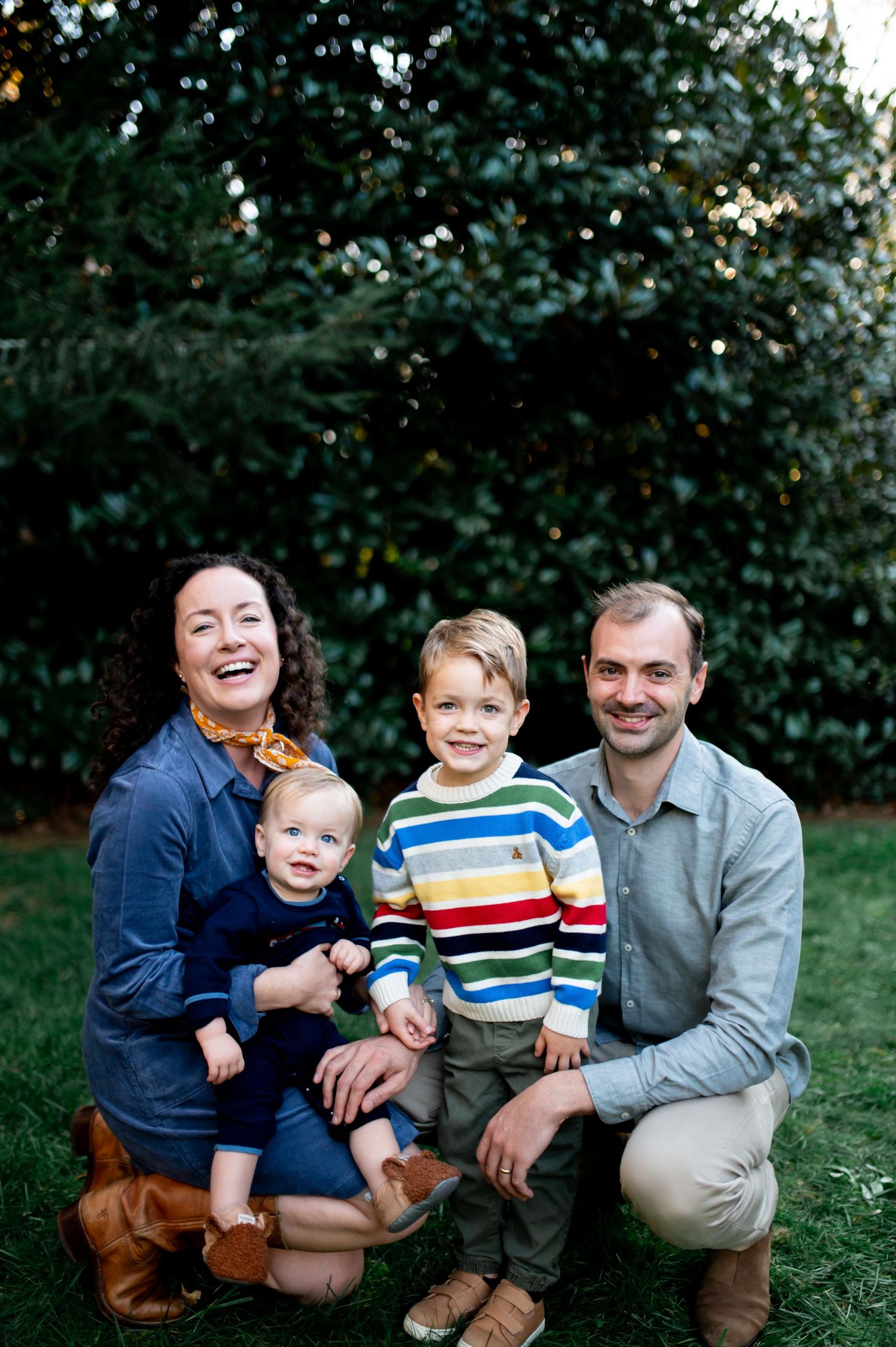 Happy family of four posing outdoors on green grass with lush trees in background.