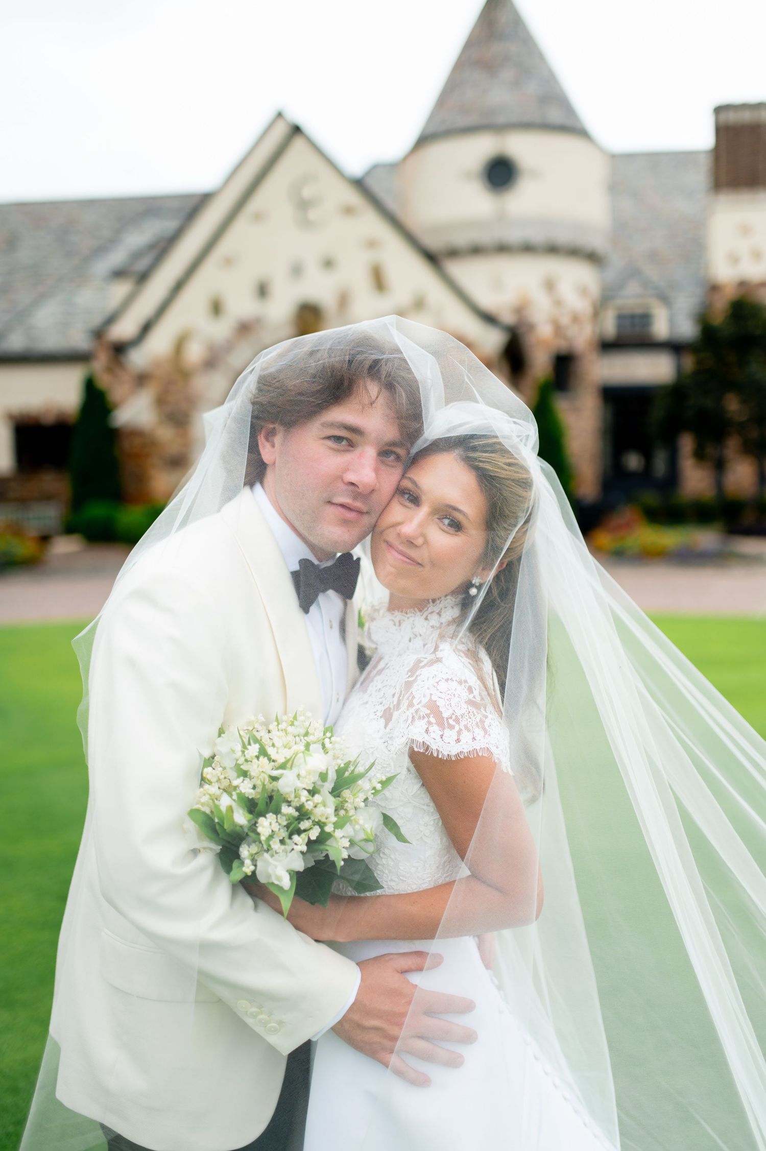 Bride and groom pose together under a flowing veil, holding a white bouquet, with a stone chapel in the background.
