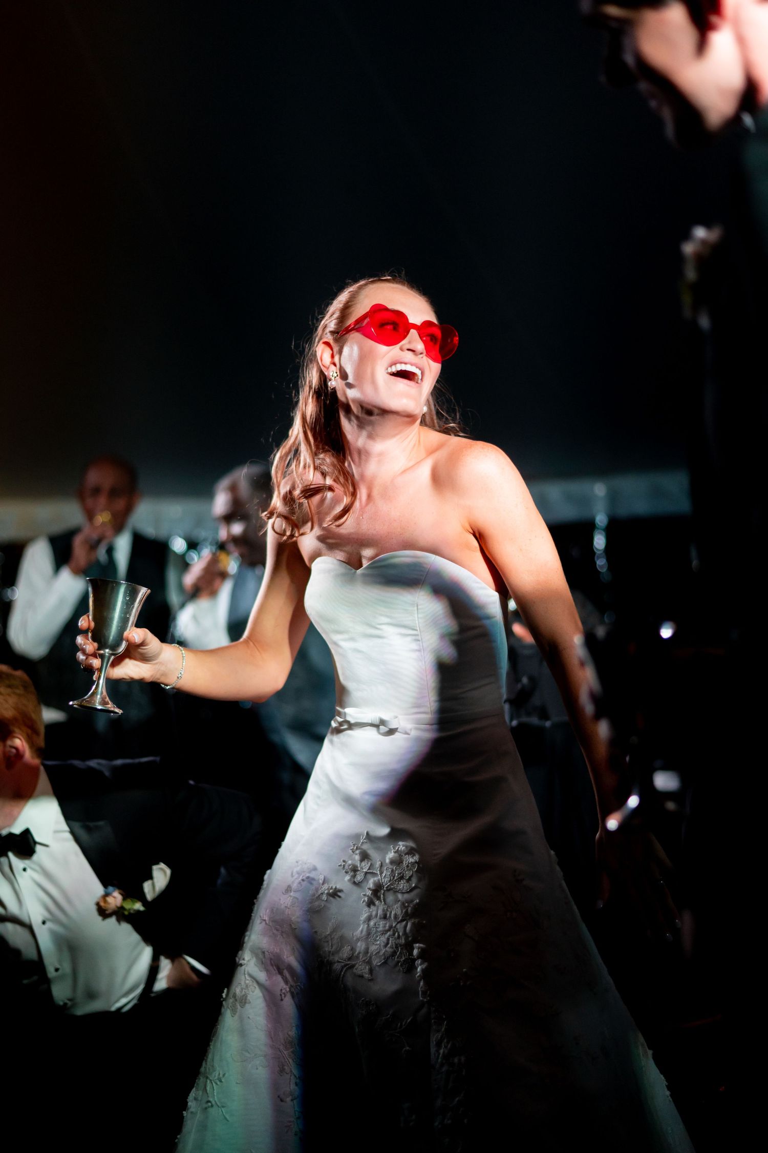 Joyful bride in white gown wearing red heart sunglasses dances with champagne at wedding reception.