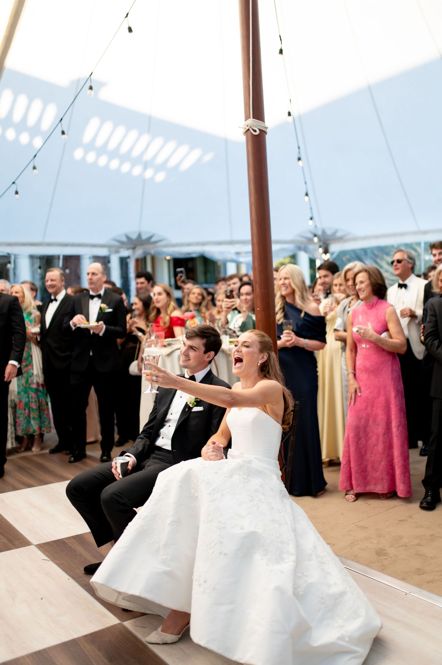 Bride and groom raise champagne glasses while seated during lively outdoor tented wedding reception.